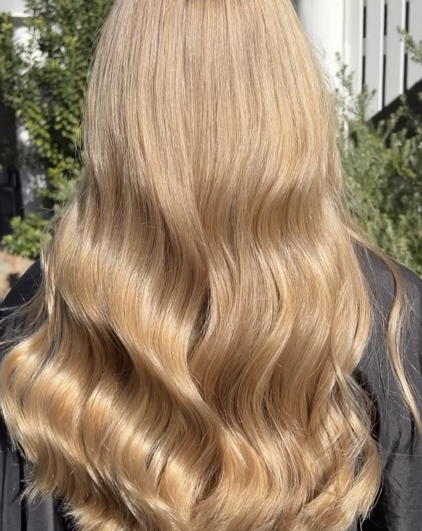 Back view of a woman with long, wavy blonde hair outdoors, with trees and a building in the background.