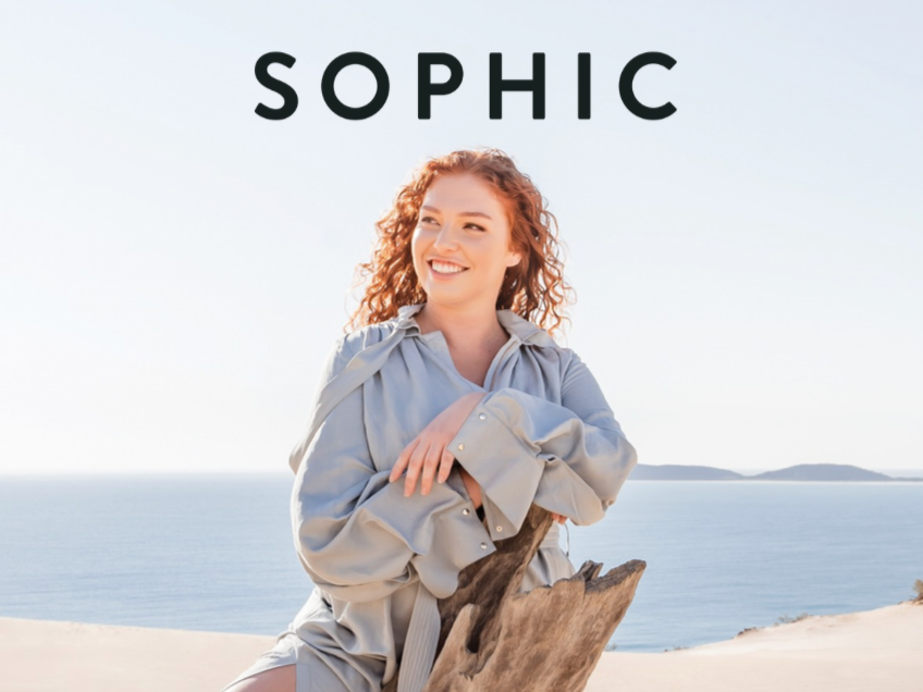 A woman with curly red hair smiling, sitting outdoors near a body of water, with the word 'SOPHIC' at the top.