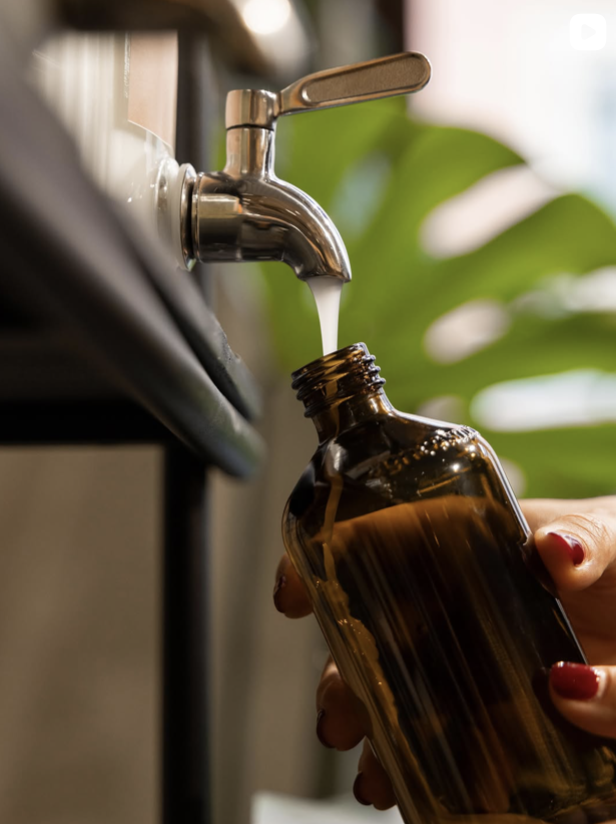 A person with red painted nails holding an amber glass bottle under a faucet dispensing liquid, with a green leafy plant in the background.