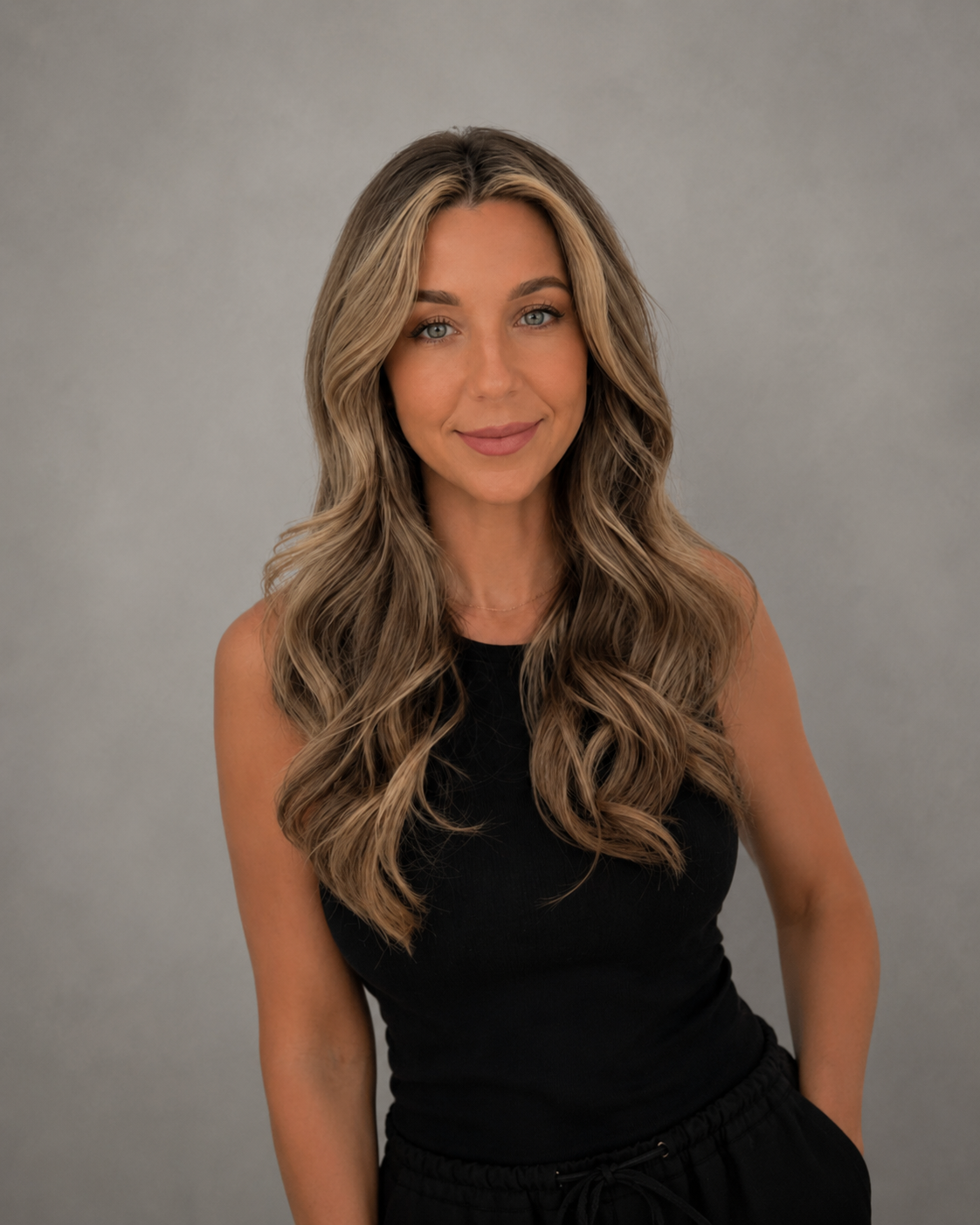 A woman with long wavy blonde hair, blue eyes, wearing a black sleeveless top, standing against a plain gray background.