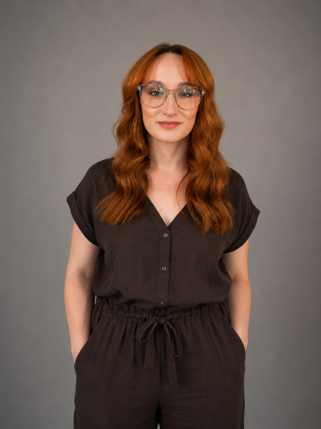 A woman with wavy red hair, glasses, and a brown outfit, standing against a gray background.