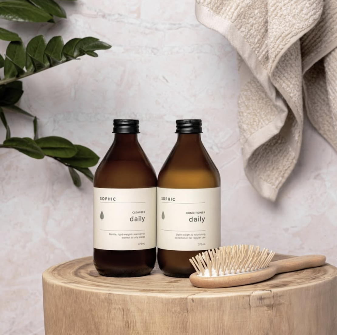 Two amber bottles labeled 'SOPHIC daily' for cleanser and conditioner on a wooden surface, with a wooden hairbrush and green leaves nearby, and a beige towel draped in the background.