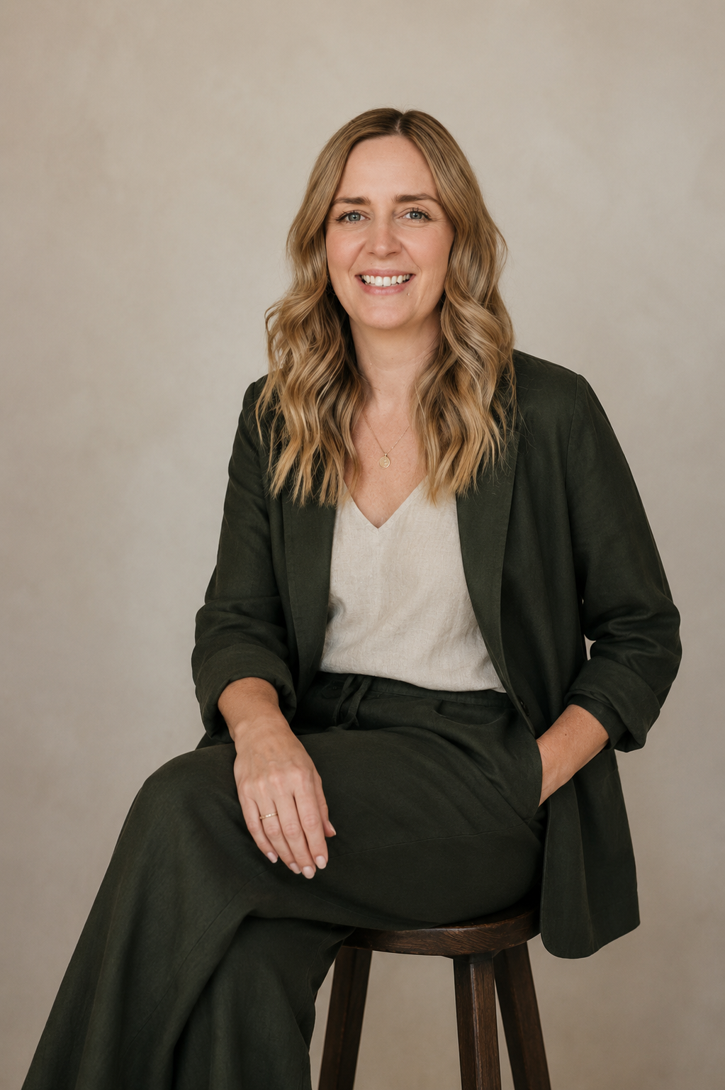 A woman with blonde wavy hair sitting on a wooden stool, wearing a dark blazer, light-colored top, and dark pants, smiling at the camera against a neutral background.