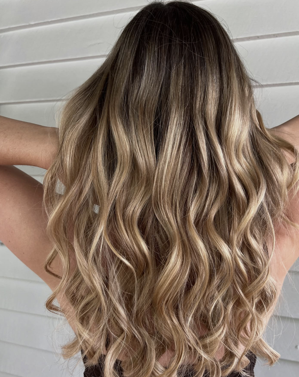 Woman with long, wavy, ombre hair in shades of blonde and light brown, standing against a white wall.