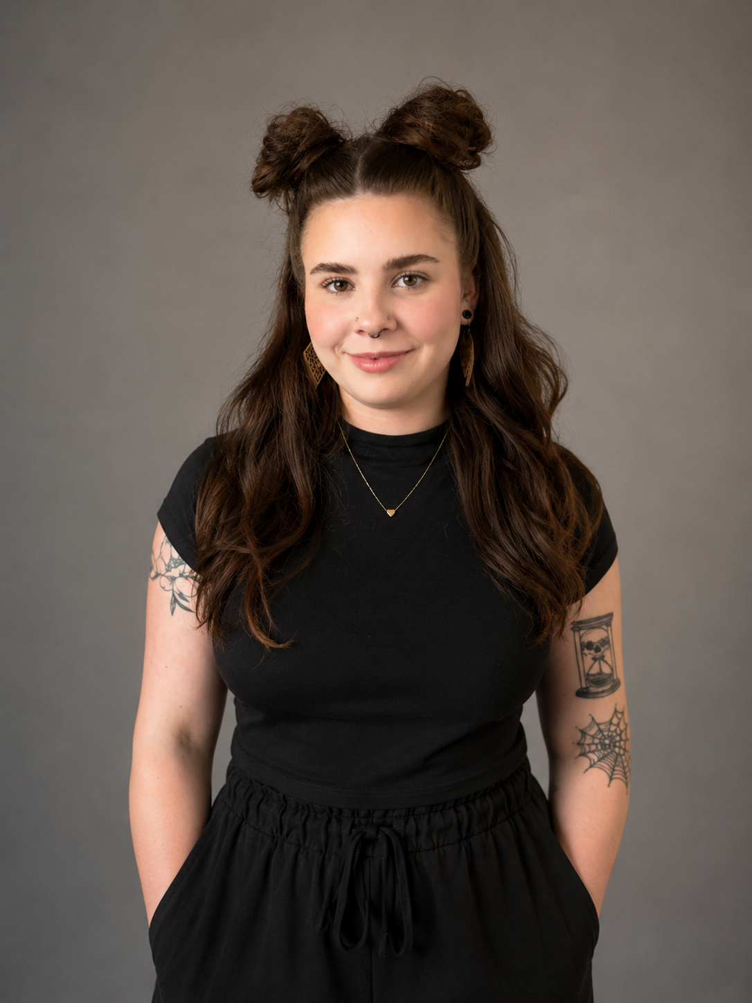 A young woman with brown hair styled in two buns, wearing a black t-shirt and black pants, standing against a gray background. She has tattoos on her arms and is smiling slightly, facing the camera.