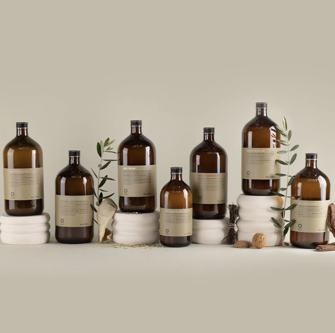 Set of amber glass bottles with labels, arranged on white ceramic bases with green foliage, nuts, and natural decor elements on a neutral background.