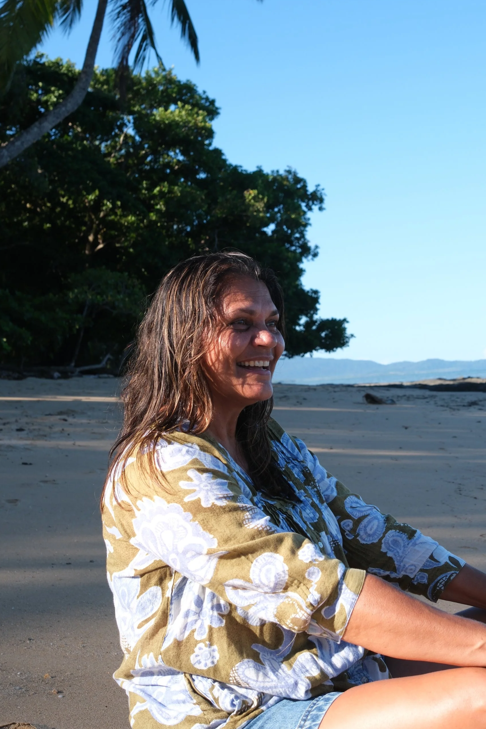 A woman with long, wavy hair sitting on a beach, smiling and enjoying the outdoors, with trees and a clear blue sky in the background.