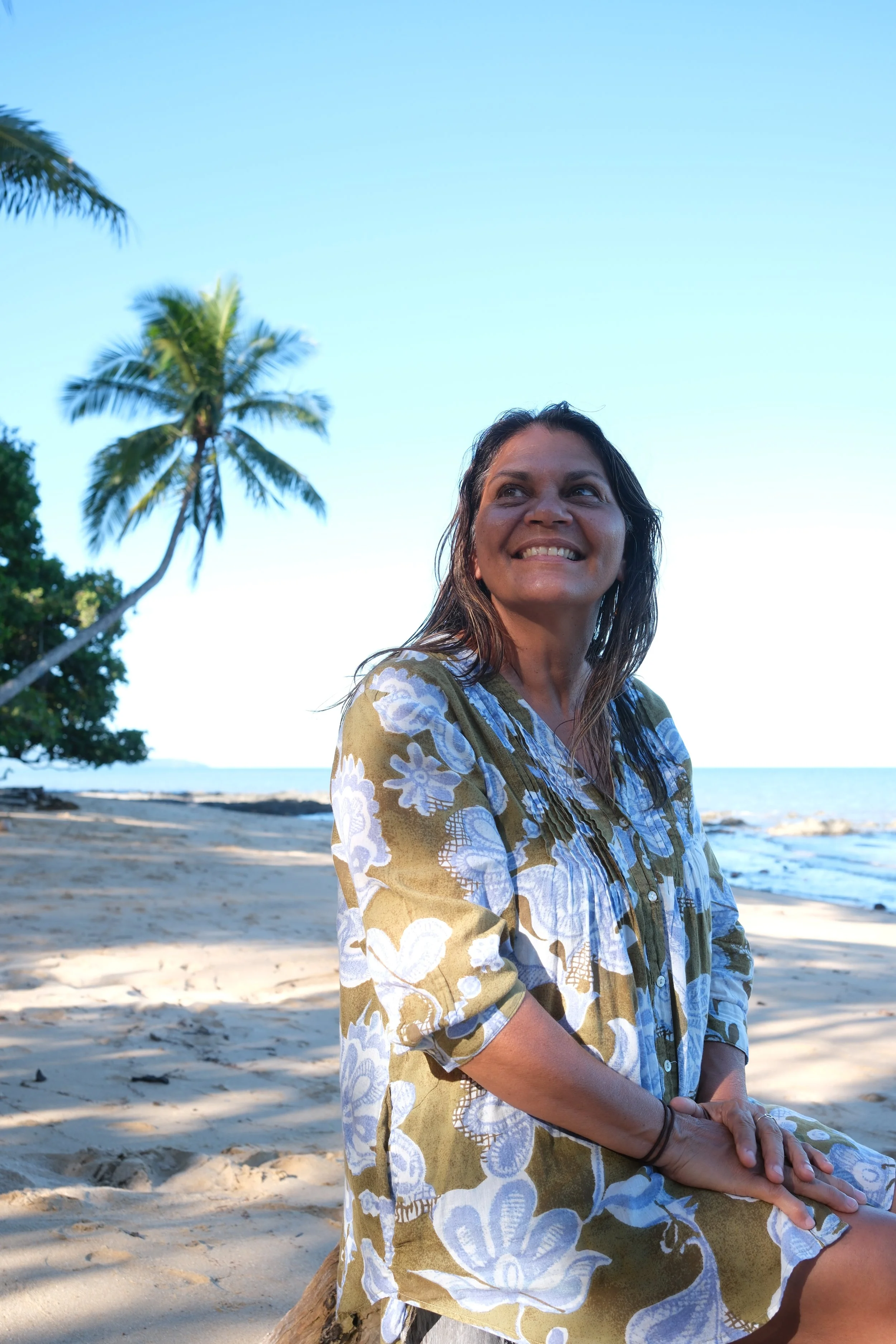 A woman with dark hair and a big smile sitting on a sandy beach, wearing a patterned yellow, white, and blue dress, with palm trees and calm ocean water in the background during daytime.