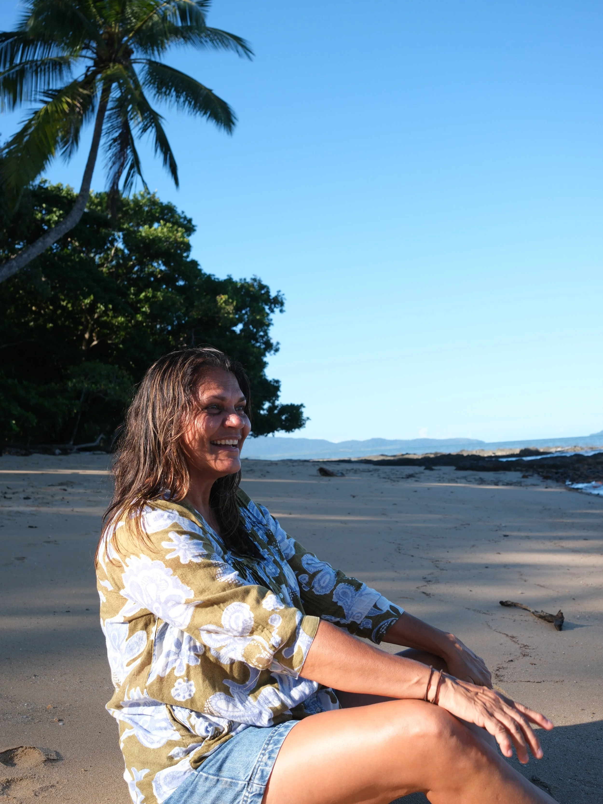 Woman sitting cross-legged on a sandy beach near the ocean, wearing a floral-patterned shirt and denim shorts, with palm trees and blue sky in the background.