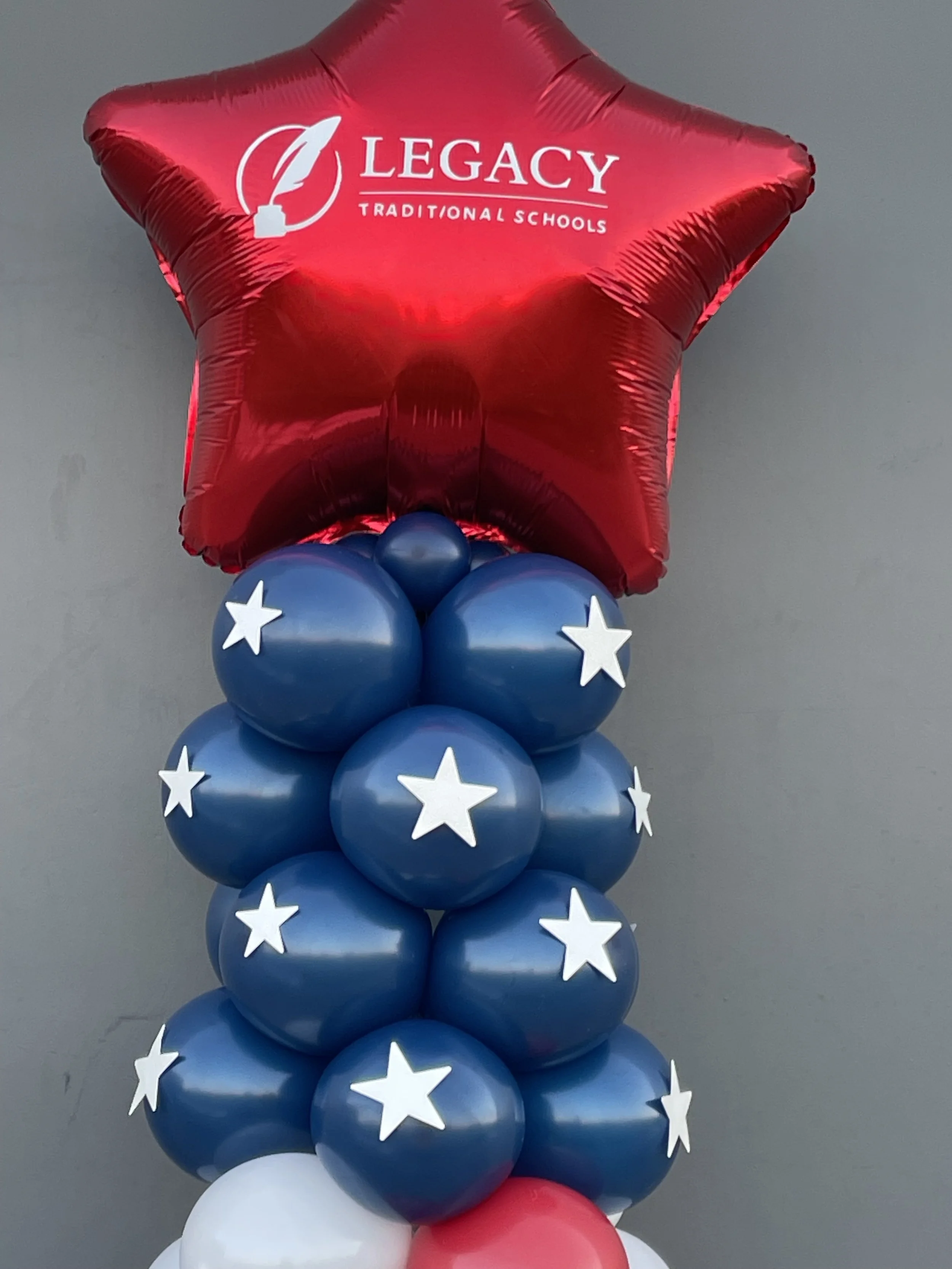 Balloon arrangement with a large red star-shaped balloon with the text "Legacy Traditional Schools" on it, surrounded by multiple blue balloons decorated with white stars and some white and red balloons at the bottom.