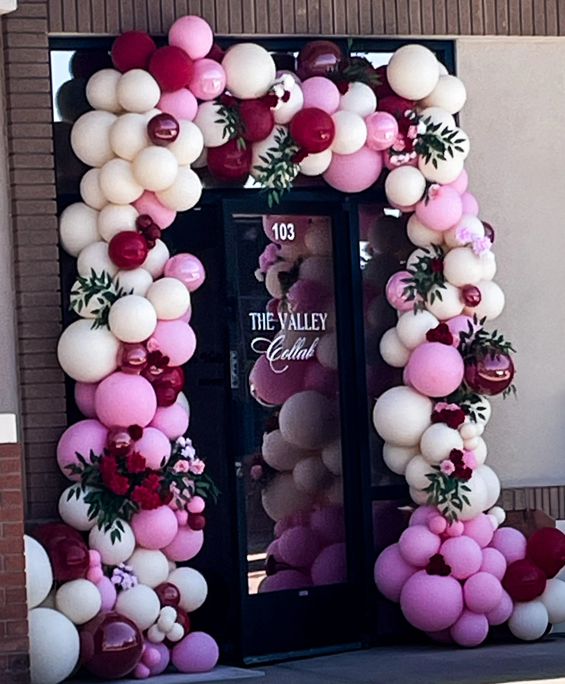 Decorative organic balloon arch with pink, white, and burgundy balloons surrounding a glass door entrance, with green leaves and flowers interspersed. 1 year anniversary 