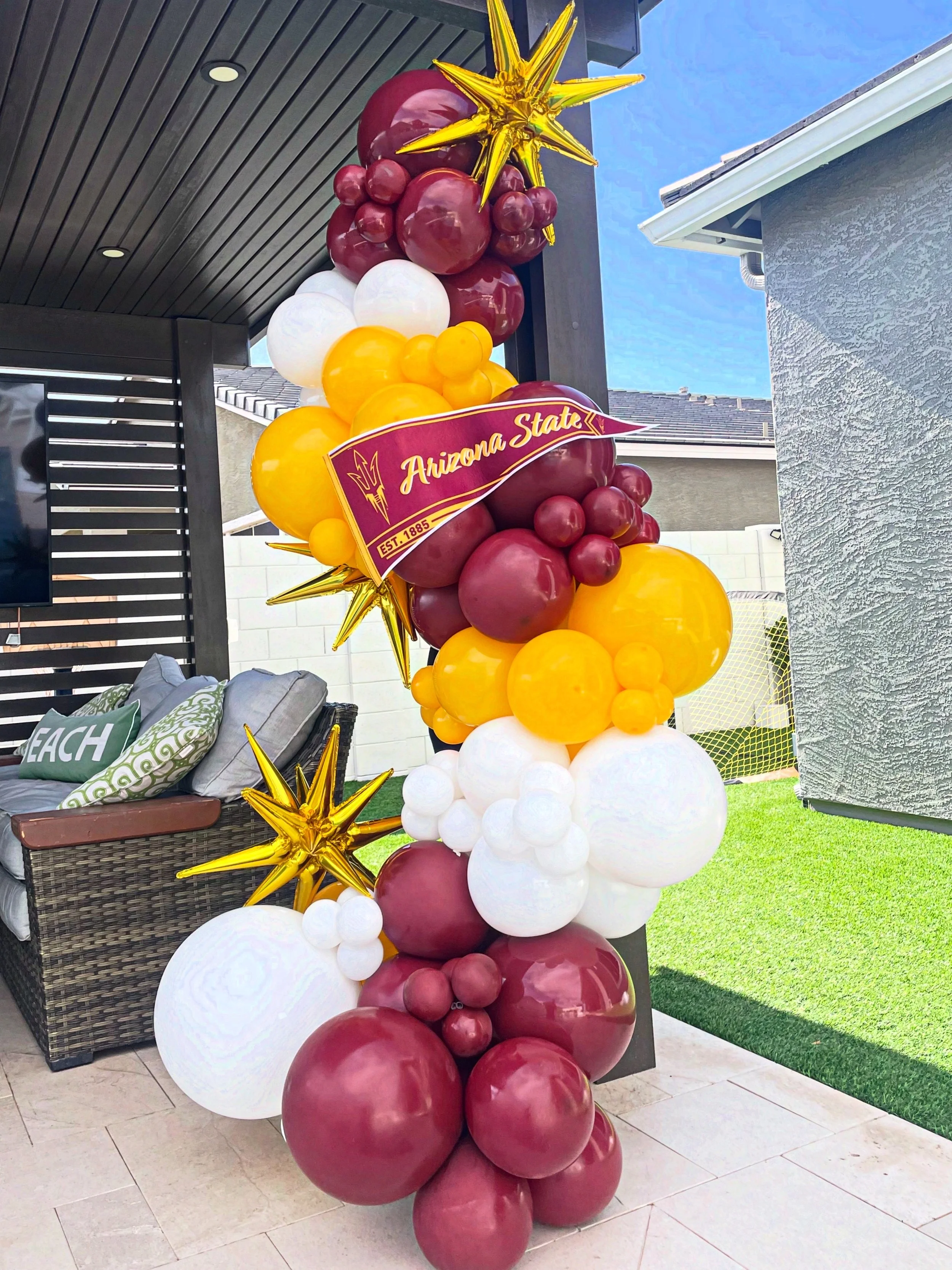 ASU Graduation Freestanding Organic Balloon Garland featuring maroon, white, and yellow balloons, with a maroon pennant that reads 'Arizona State' and includes the established date '1885', accented with gold star-shaped balloons, set on a patio 