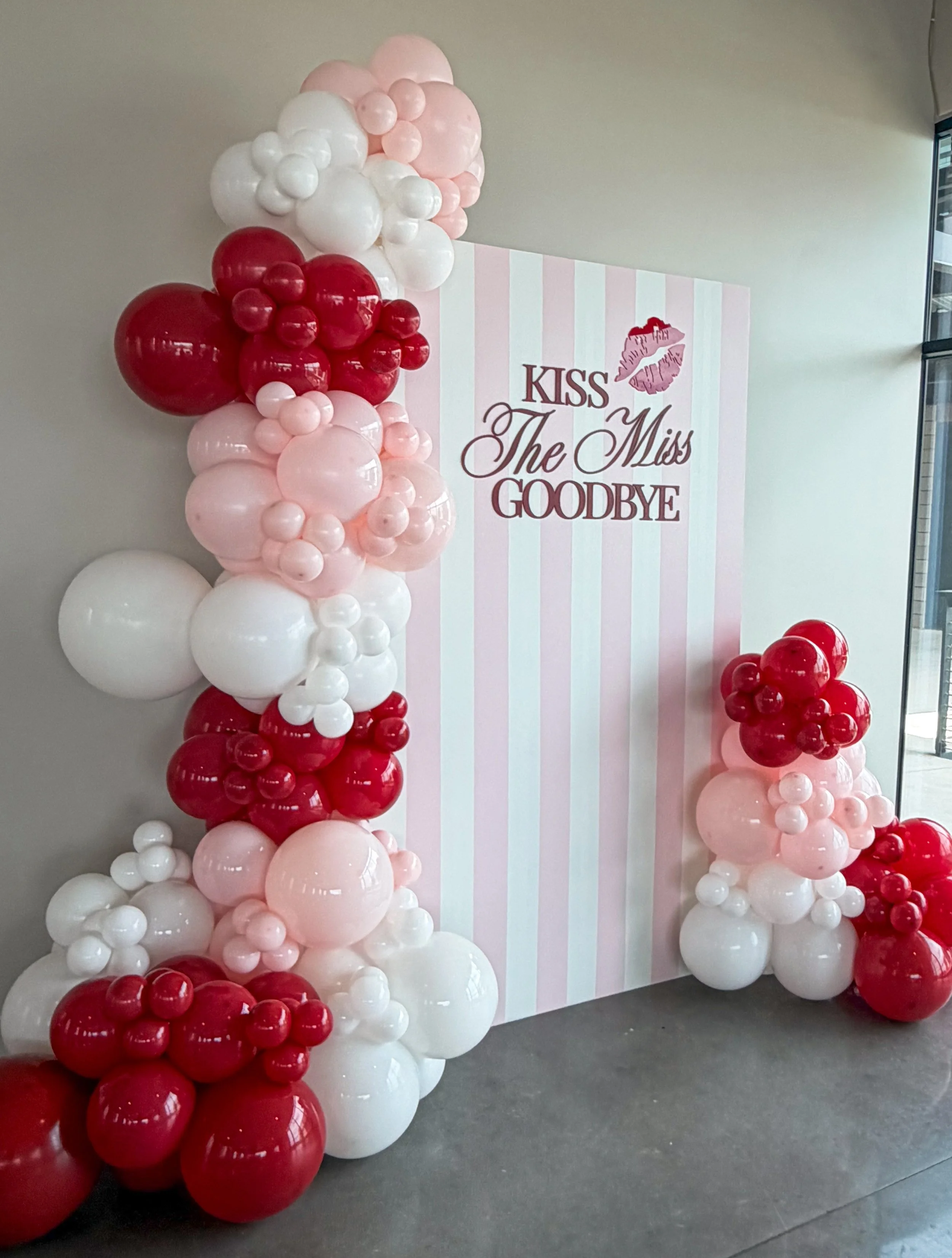 Decorative balloon arrangement with pink, white, and red organic balloon garland in front of a pink and white striped backdrop. The backdrop features a quote "kiss the miss goodbye".  Bridal Shower