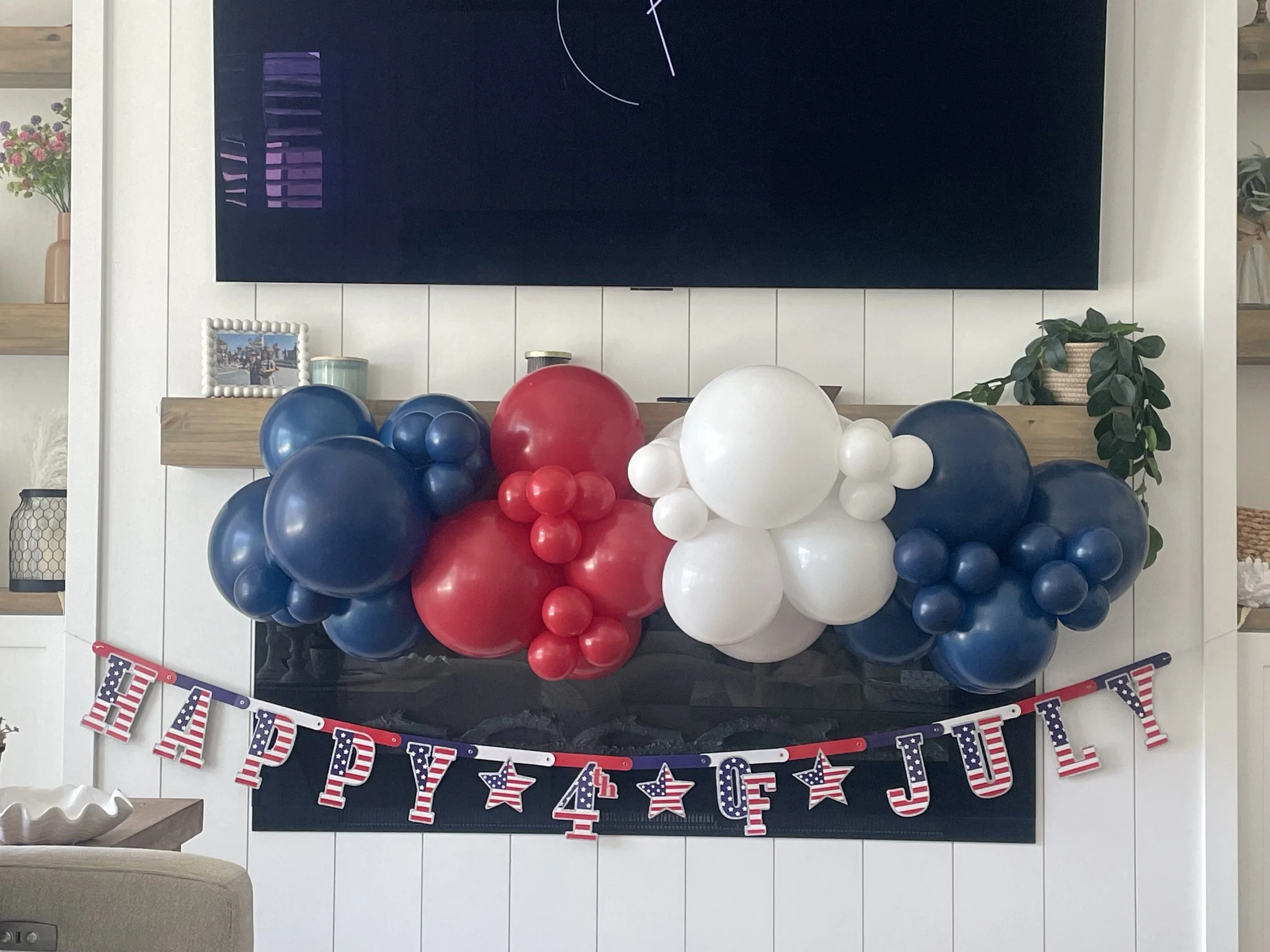 Red, white, and blue balloons arranged in front of a dark fireplace with a holiday-themed banner that says "HAPPY the 4th OF JULY" with stars and American flag patterns.