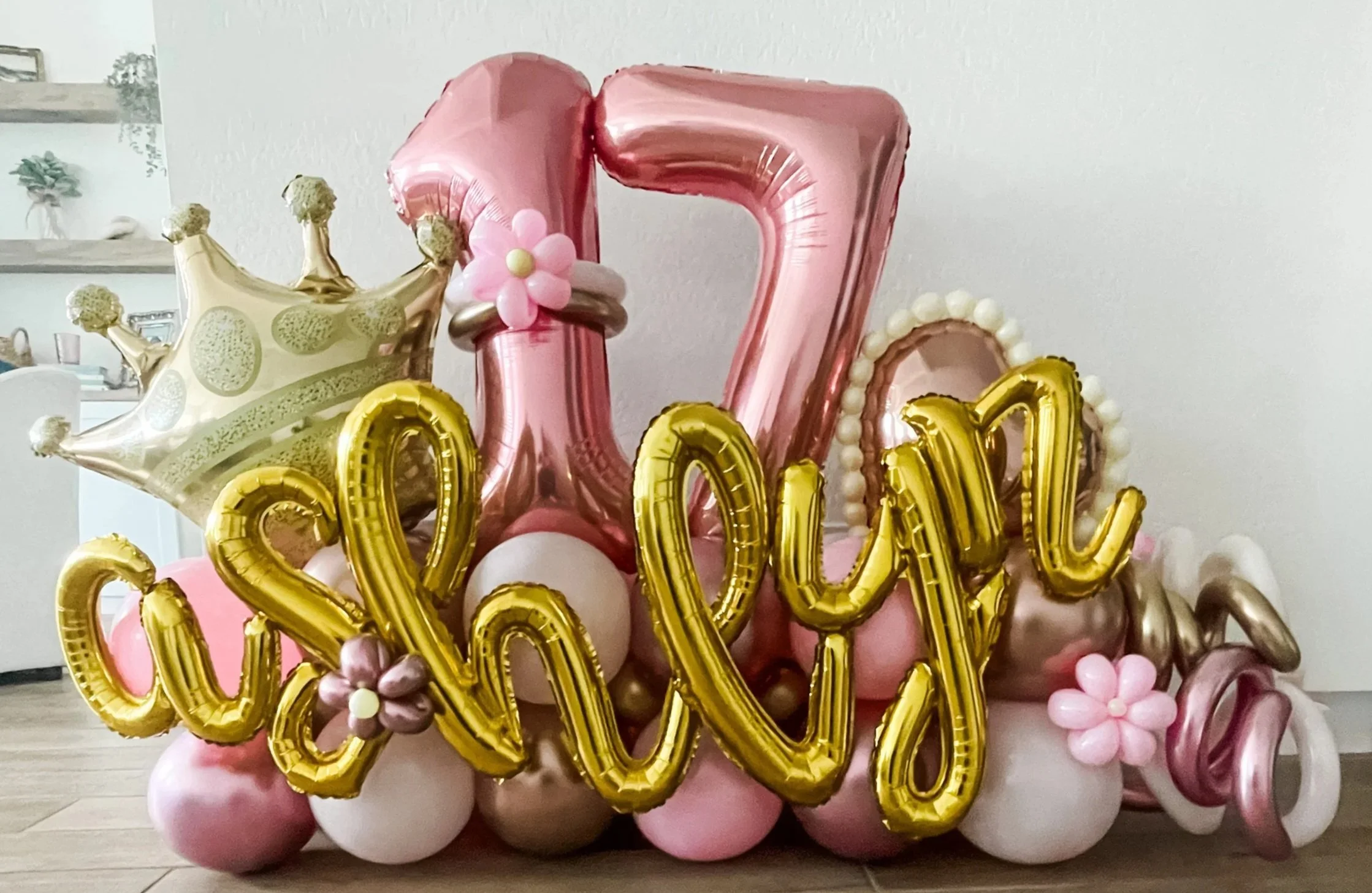 Balloon display with pink, white, and gold balloons spelling out 'love' and a gold crown balloon on top, decorated with pink and gold flowers.