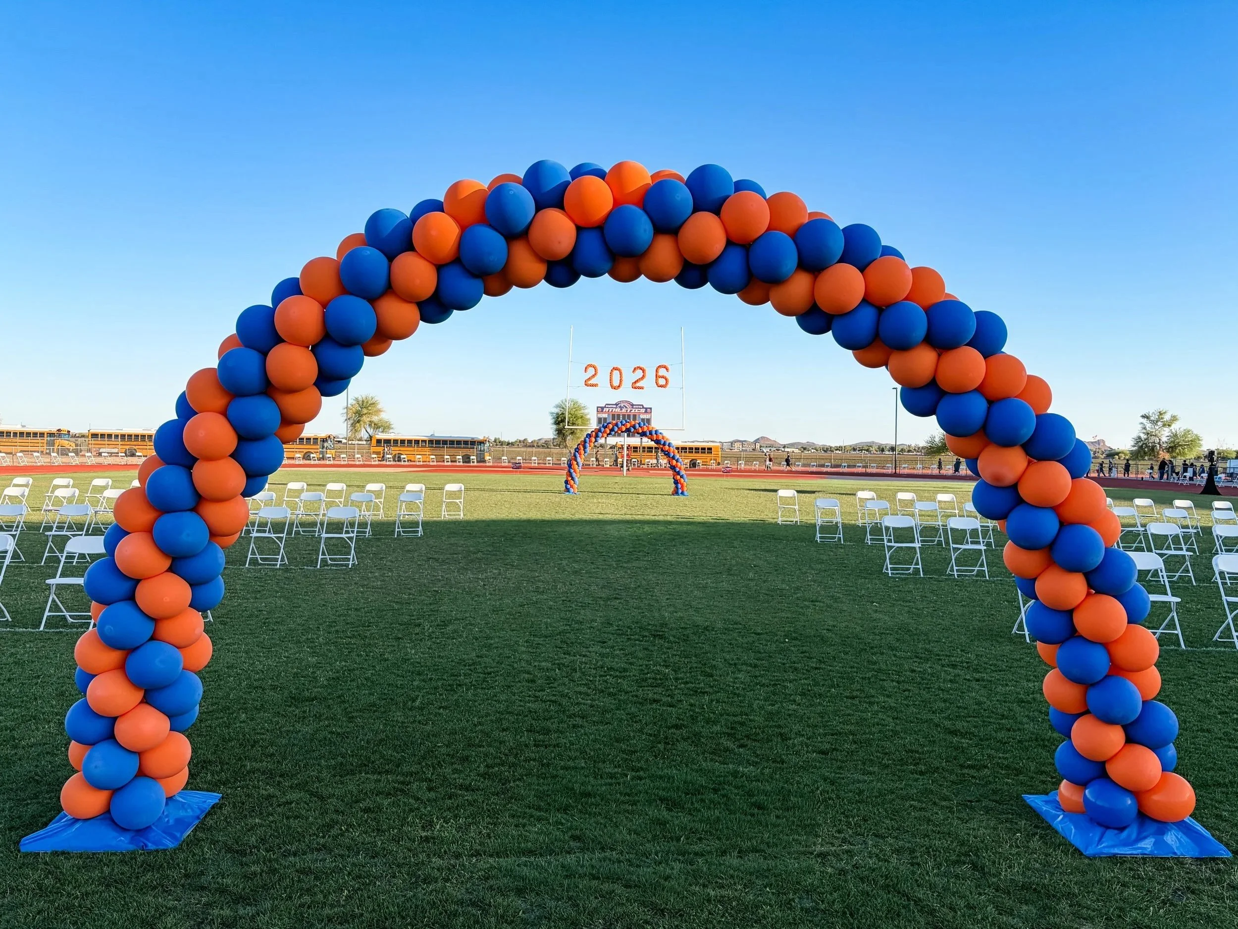 A large arch made of blue and orange balloons set up on a sports field with seating and festive decorations in the background and the numbers '2026' hanging in the distance.
