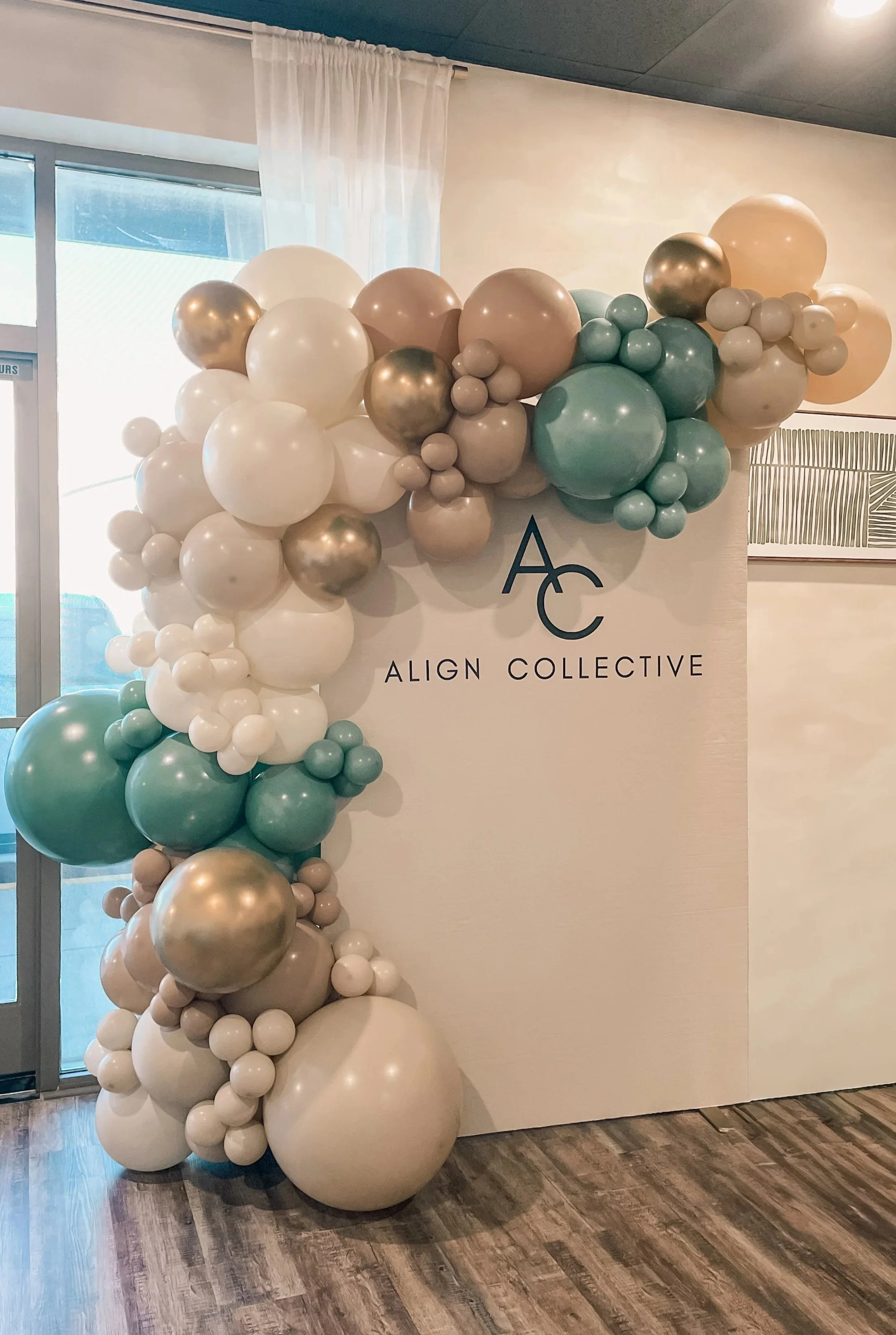 Decorative balloon arrangement with white, cream, teal, and metallic gold balloons next to a wall with the logo 'Align Collective' on it.
