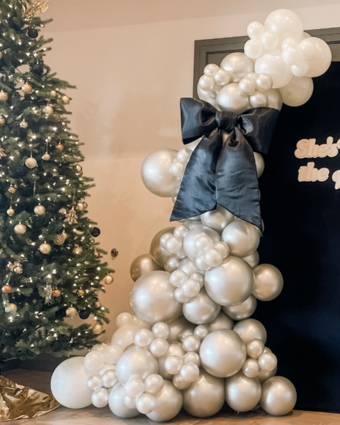 Decorative Christmas display with a large cluster of white and silver balloons atopped with a black bow, next to a decorated Christmas tree with gold and silver ornaments.