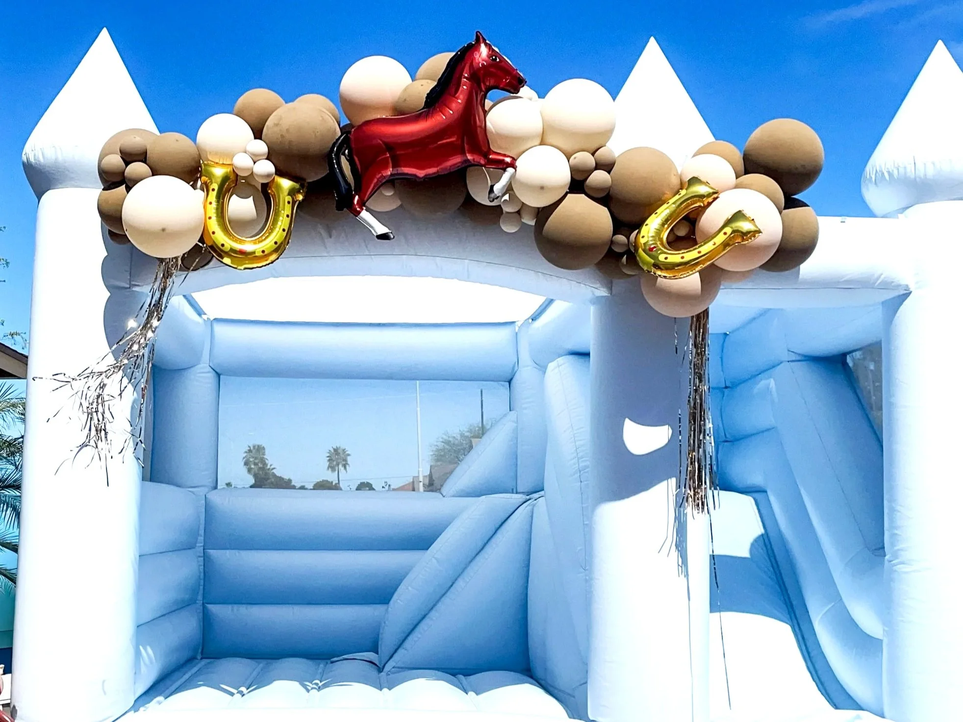 A white inflatable bounce house decorated with balloons, featuring a red and black horse-shaped balloon as a centerpiece. The bounce house has a castle theme, with white castle towers visible on both sides, and is set outdoors against a bright blue s