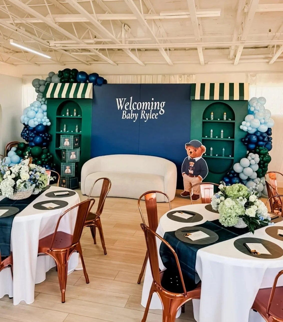 Baby shower celebration setup with a backdrop that says "Welcoming Baby Rylee," decorated with balloons in shades of blue, white, and green, along with a white couch, floral centerpieces, and tables arranged for guests.