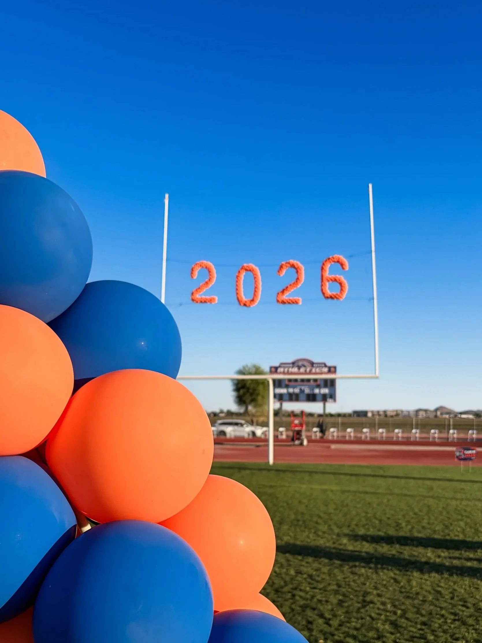 A cluster of blue and orange balloons in the foreground, with a football field and goalpost in the background. A large '2026' sign made of balloons is hung across the goalpost.
