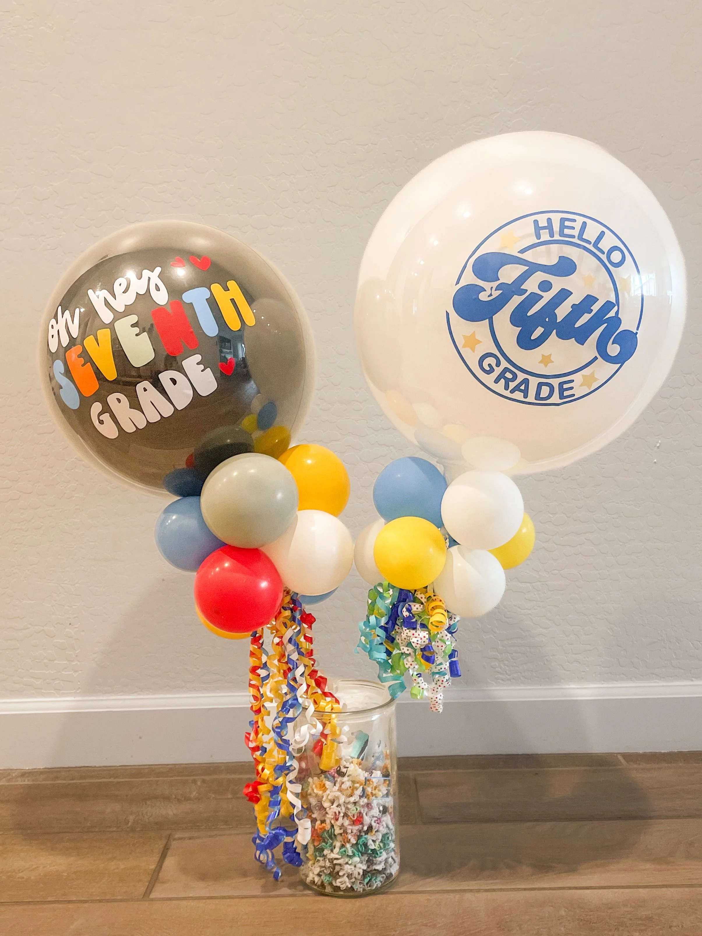 A tall glass jar filled with colorful paper shreds, topped with balloons and two large balloons with decorative prints. The one on the left says "Oh hey, seventh grade" in playful colors and font, and the one on the right says "Hello Fixin'" in blue 