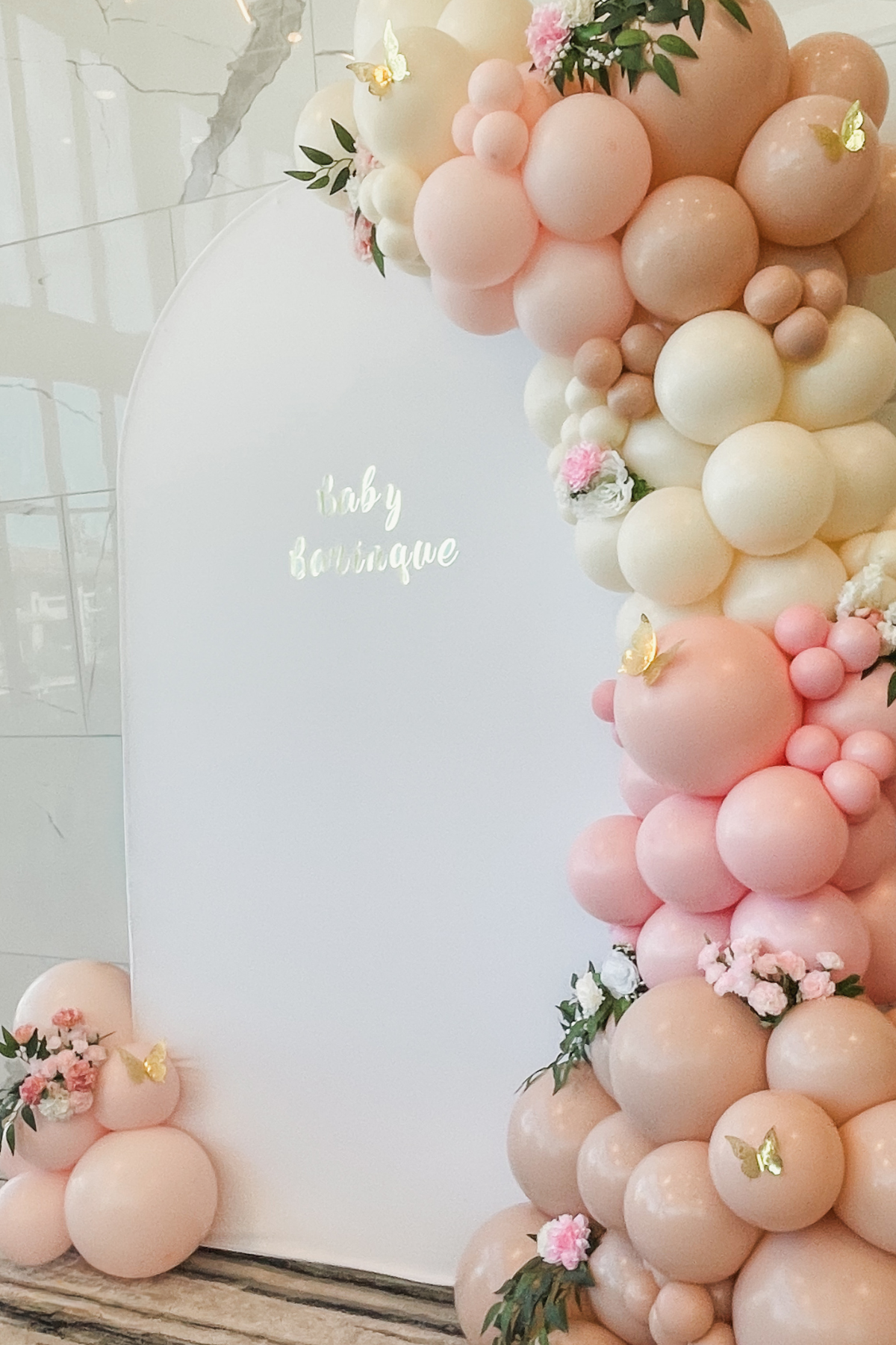 A decorative display of pink, peach, and white balloons arranged around a white board that reads 'baby boutique' with some flowers, greenery, and small golden butterfly accents.