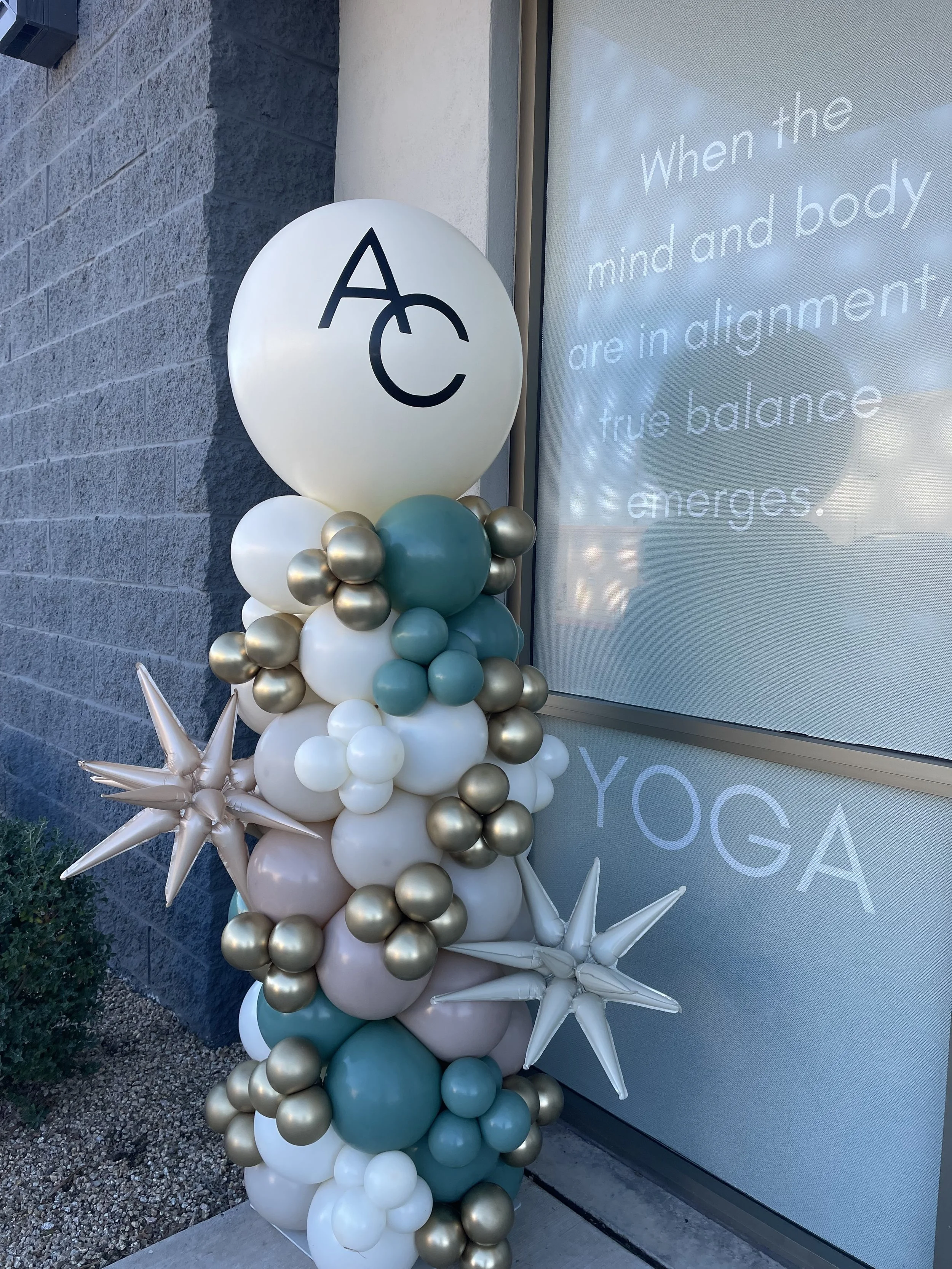 An organic balloon column with sage, teal, and gold balloons, featuring large star-shaped balloons, in front of a building. Grand Opening Balloons