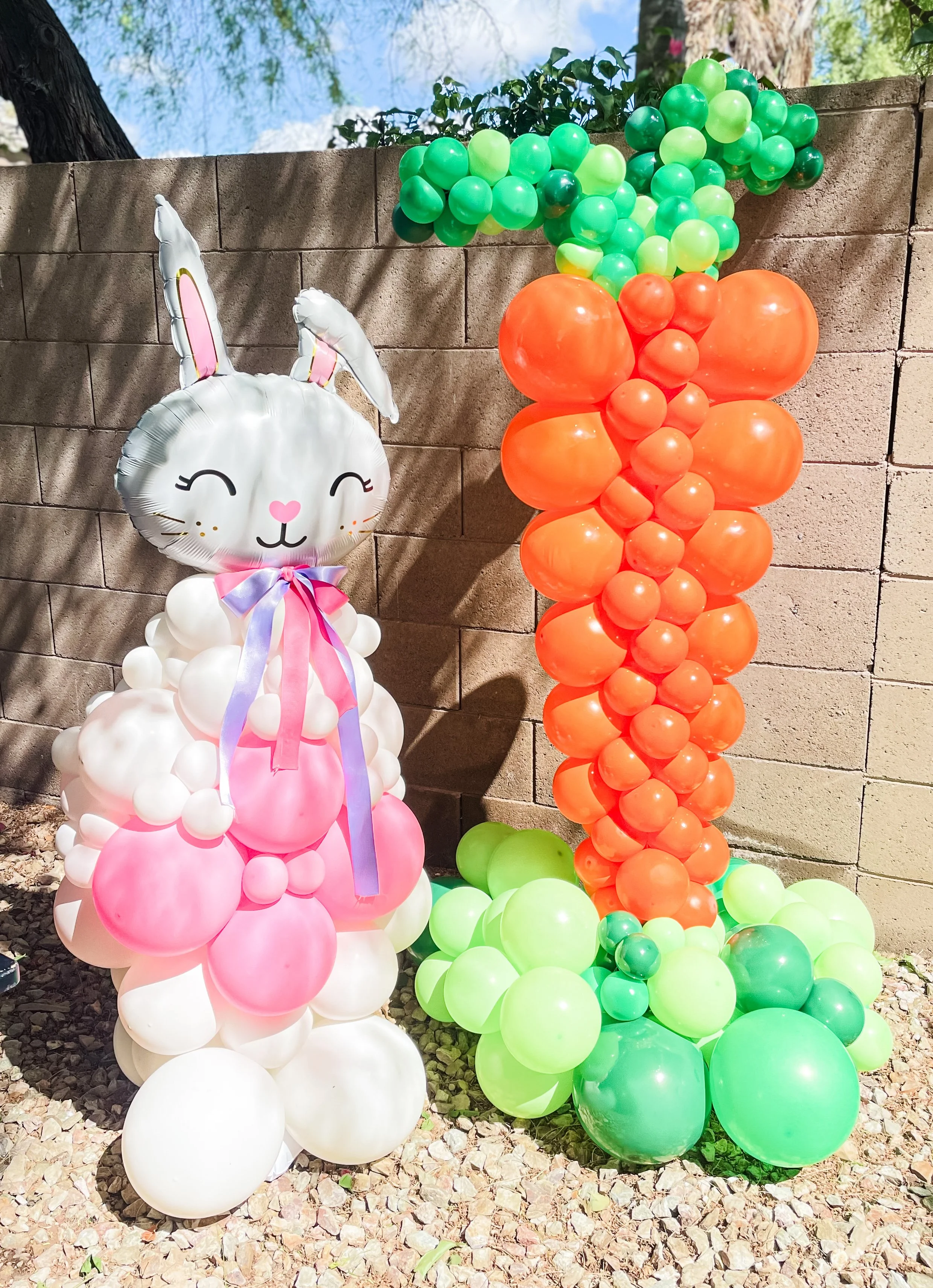 Decorative balloon display with a bunny-shaped balloon, pink and white balloons, and a green and orange balloon column outdoors against a brick wall, with trees and a blue sky in the background.