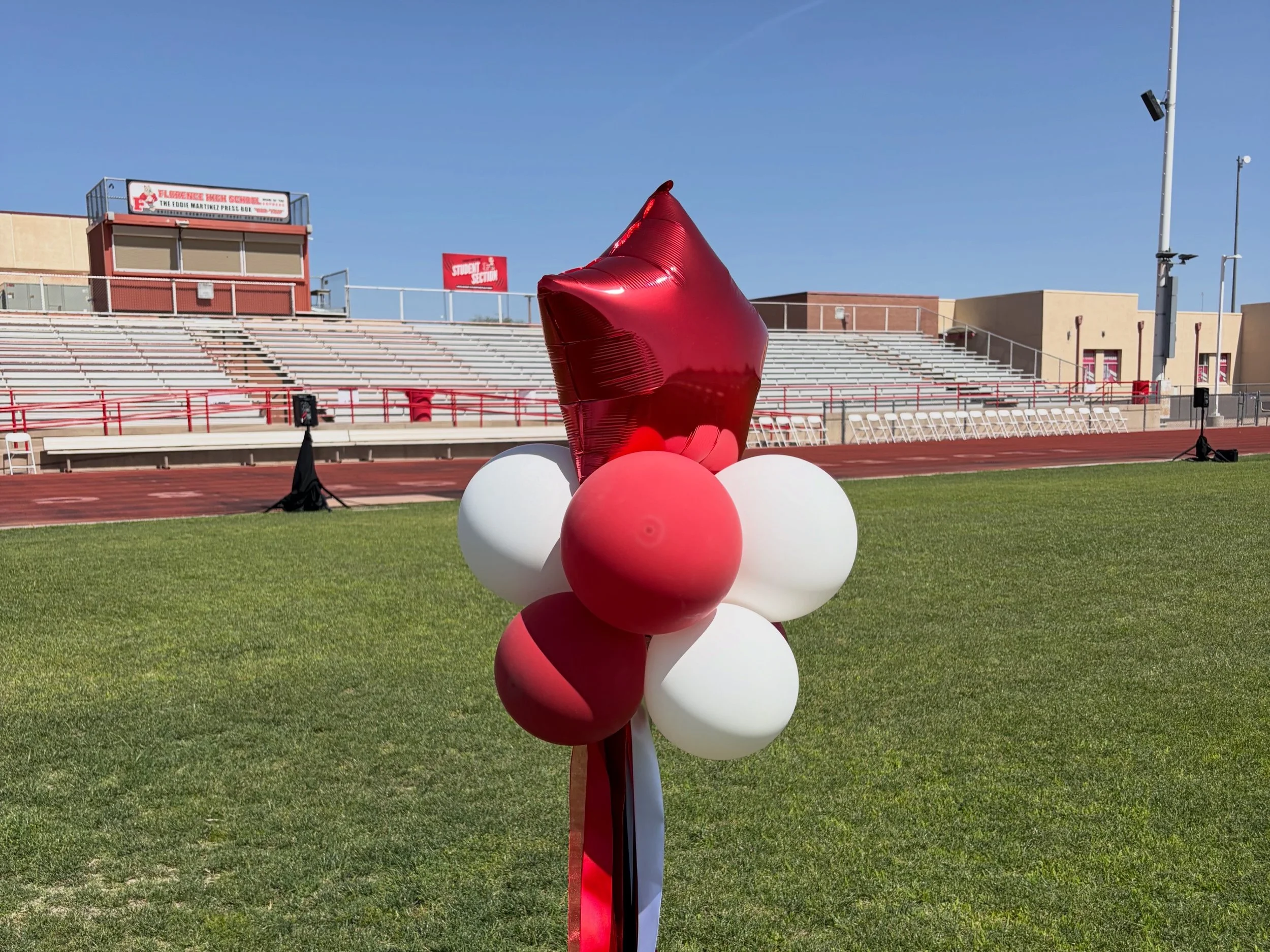 A cluster of red, pink, and white balloons tied together, set on a grassy field at a stadium with bleachers and a school sign in the background.