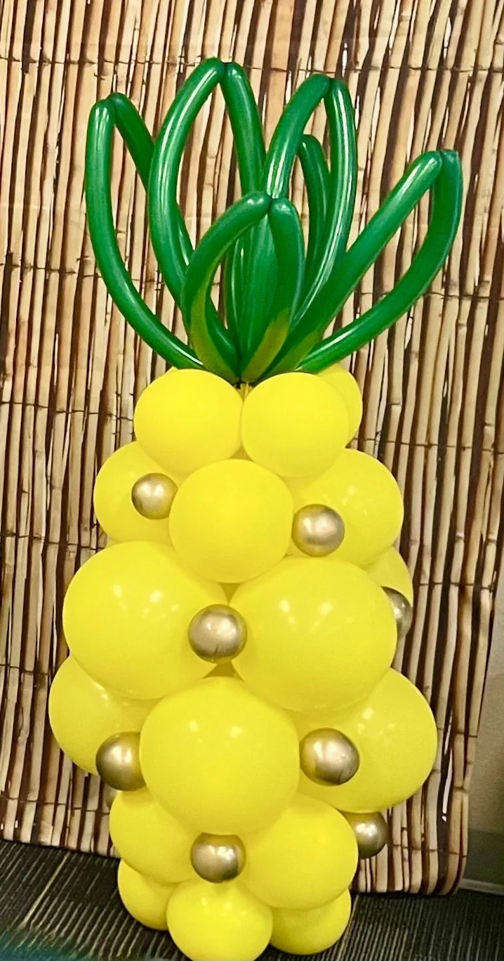 Balloon sculpture resembling a pineapple, with yellow balloons for the body, green balloon leaves at the top, and gold balloons as accents, set against a bamboo background.
