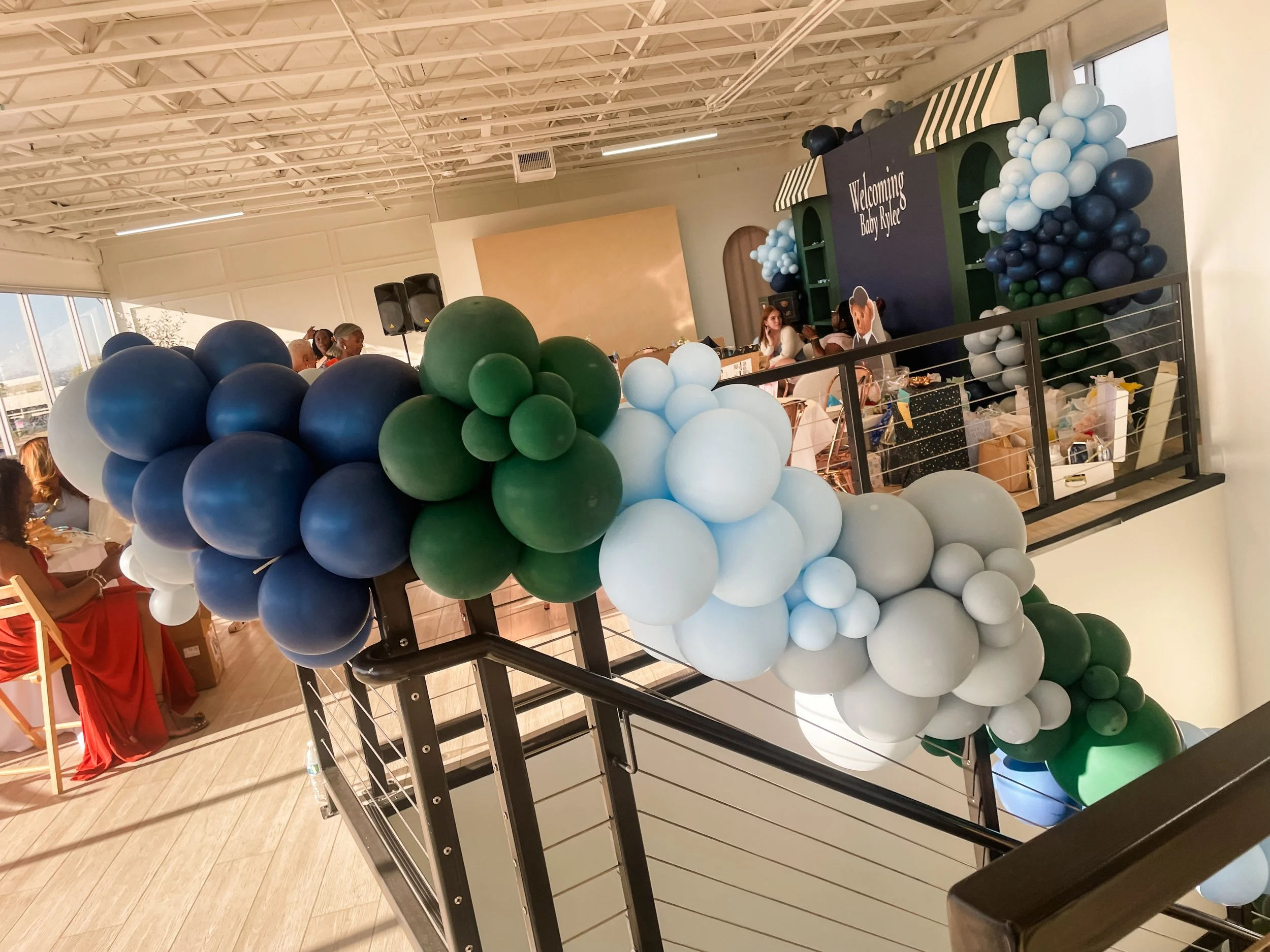 Decorations for a baby shower, featuring a large cluster of blue, green, and white balloons on a black staircase rails.  Baby Shower 