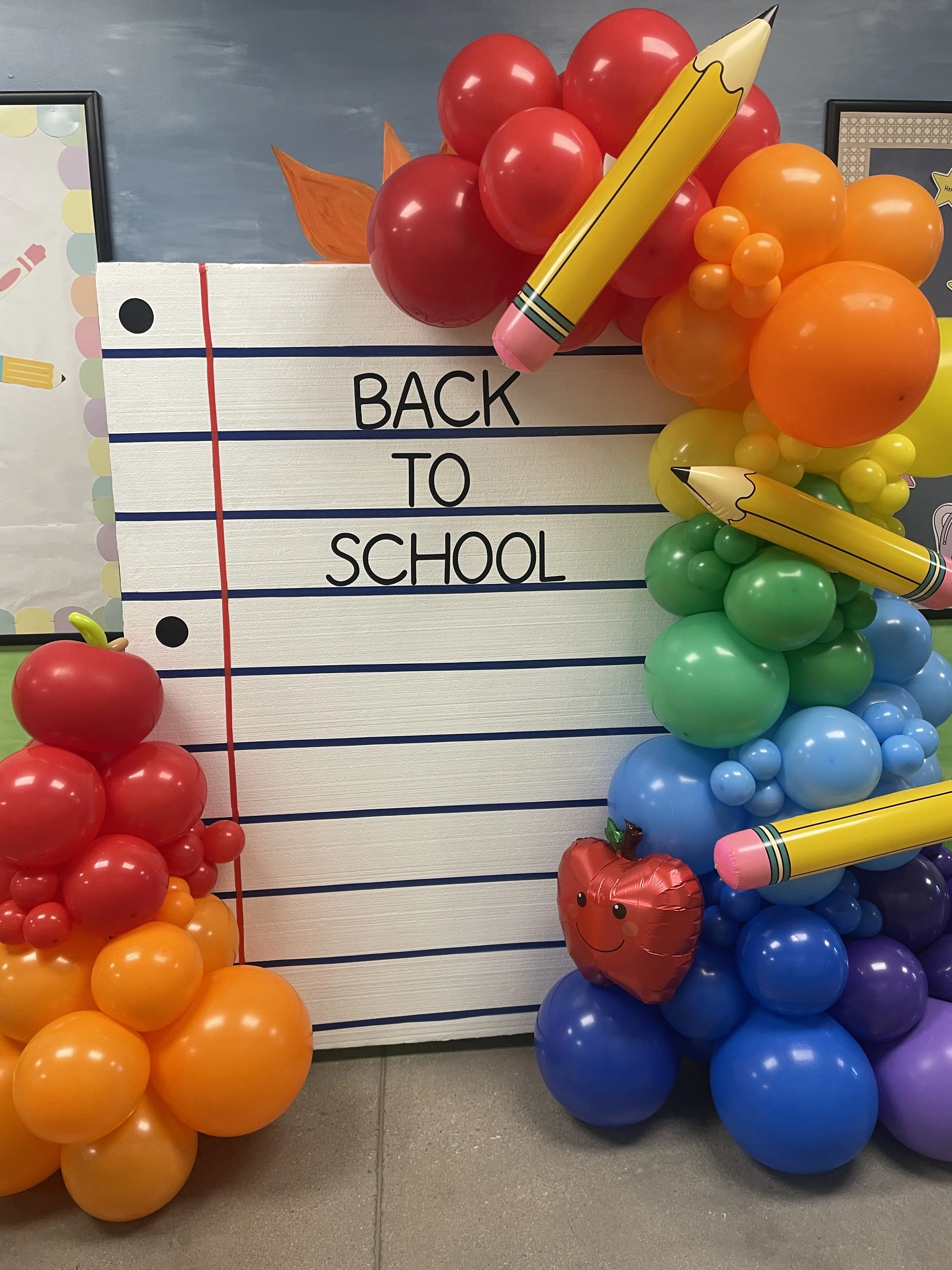 Colorful balloon display with a 'Back to School' sign, giant pencils, and apple-shaped balloons, set against a blue wall.