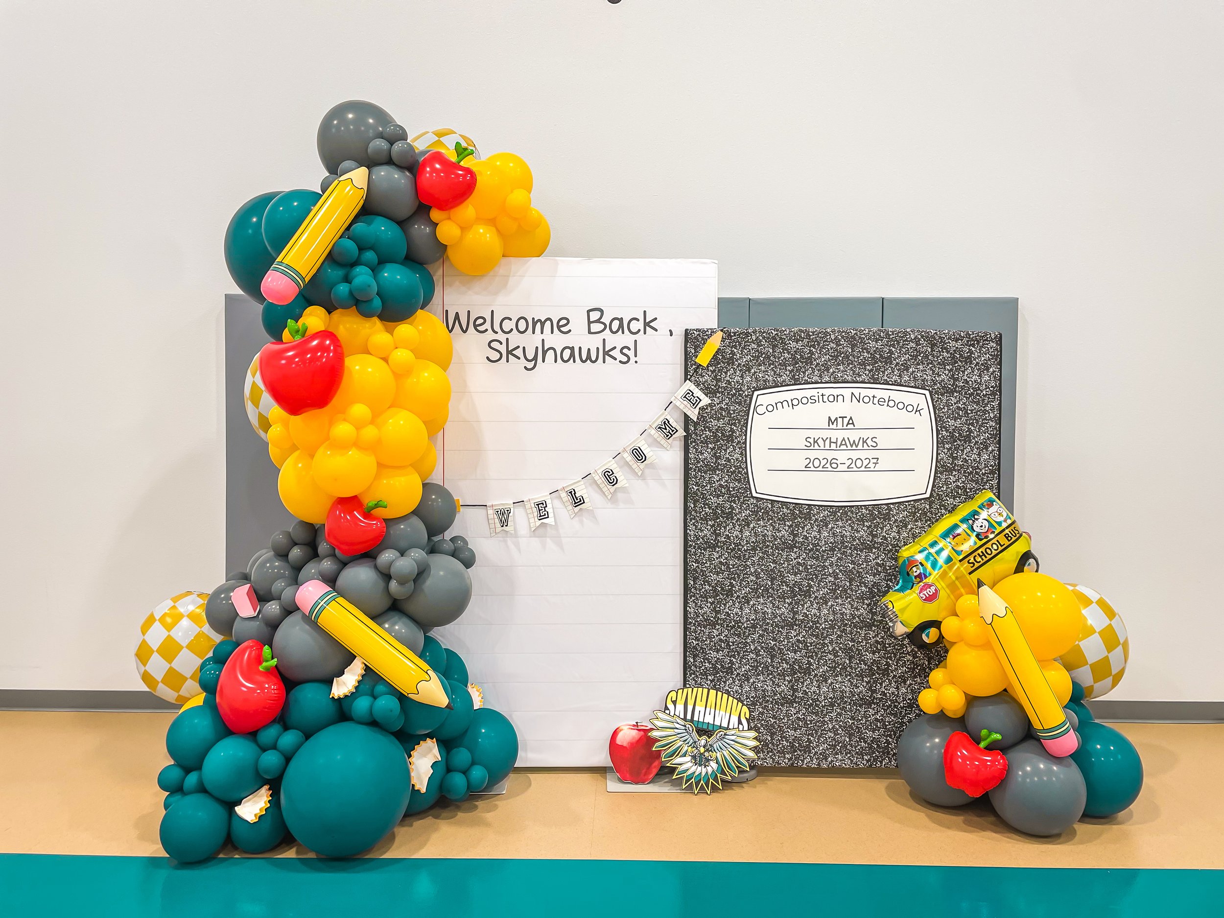 Decorative balloon arrangement with balloons in teal, gray, yellow, and red, featuring giant pencil and apple balloon shapes. A whiteboard with a 'Welcome Back Skyhawks!' message and a black and white composition notebook are also present, alongside 
