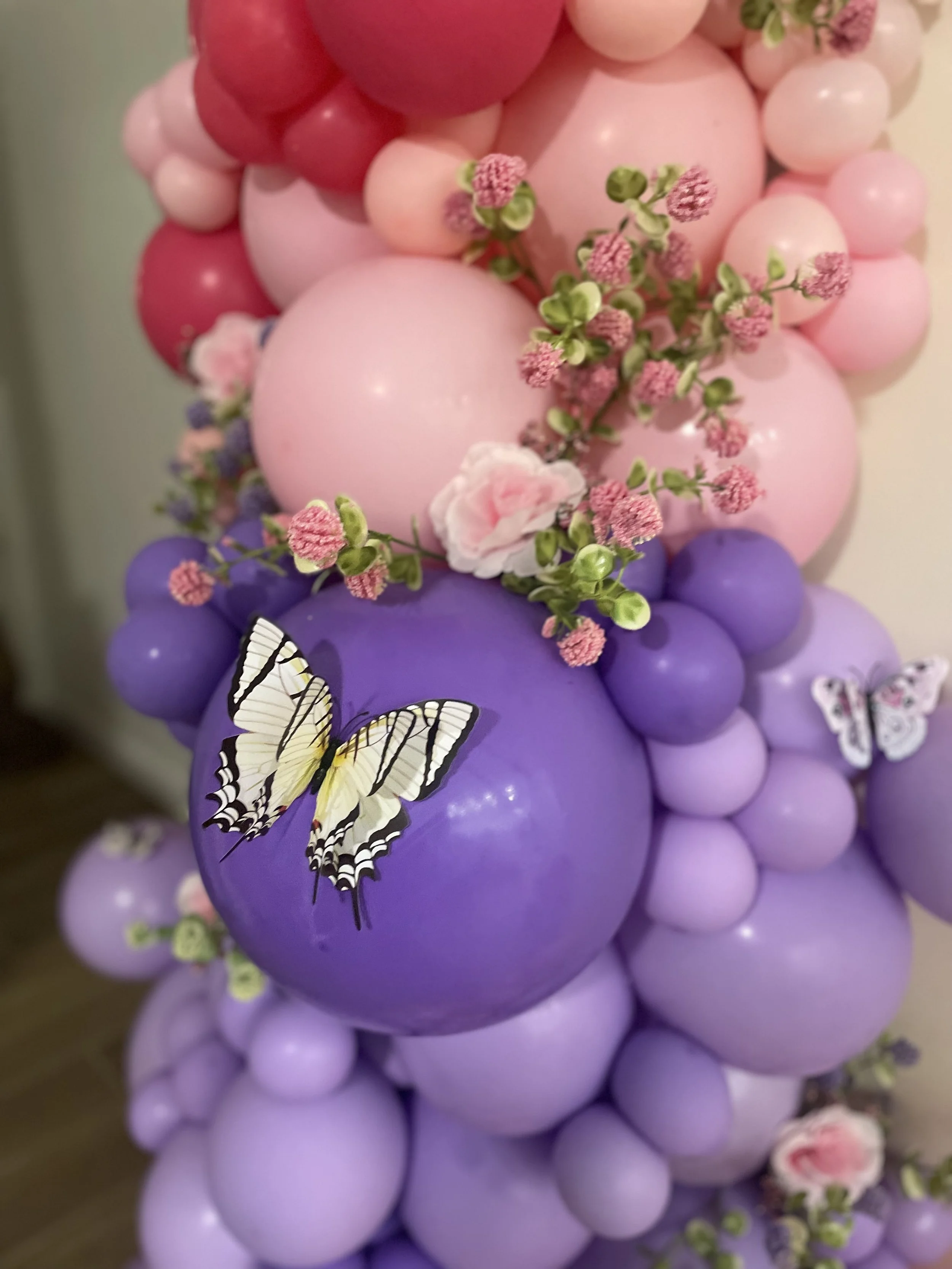 Close-up of a decorative arrangement of pink, purple, and red balloons with pink and purple artificial flowers and butterflies.