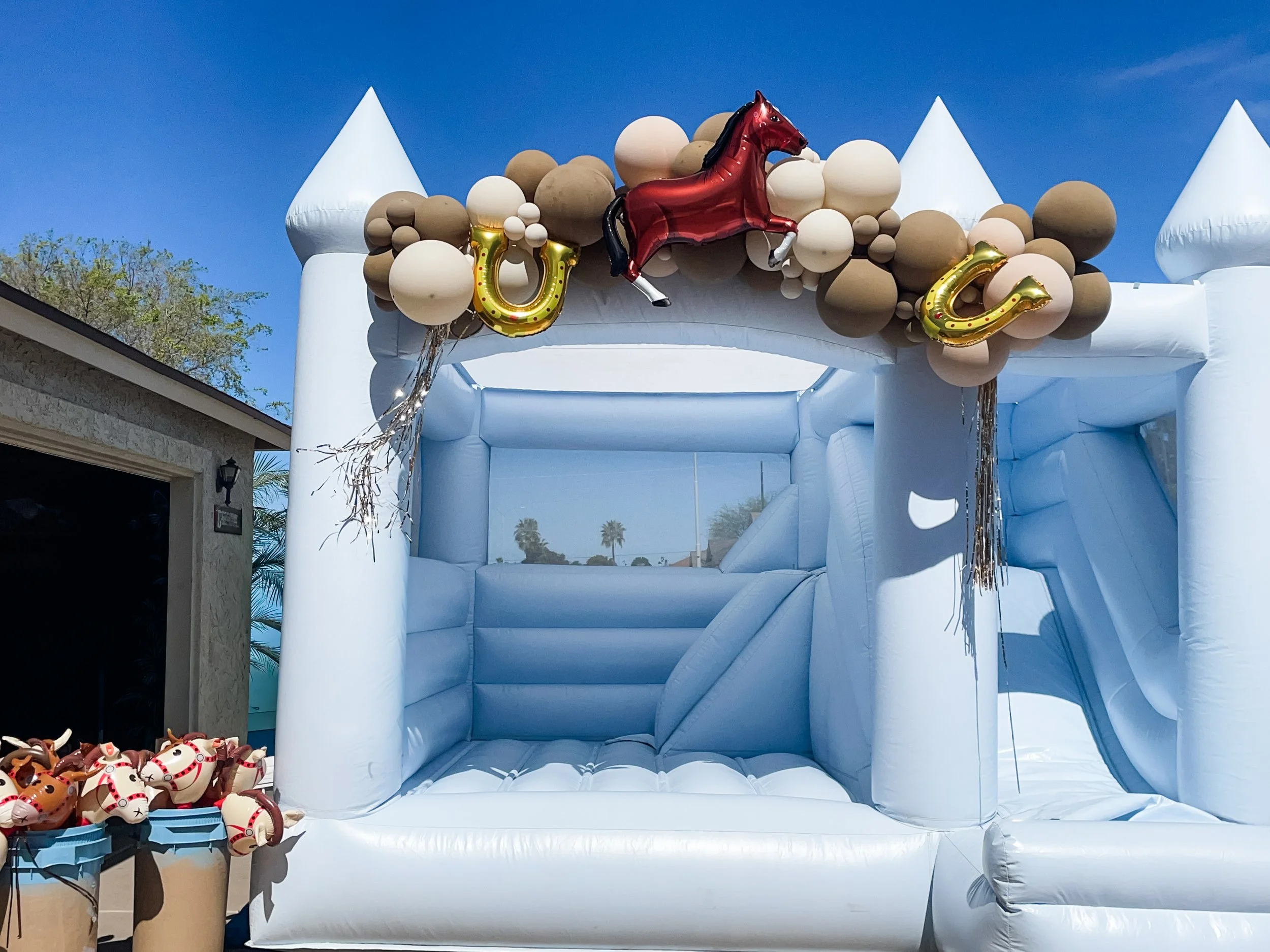 Inflatable bounce house decorated with a cluster of beige and white balloons, a red horse-shaped balloon, and gold balloon letters spelling 'U'.  Cowboy themed birthday party. 