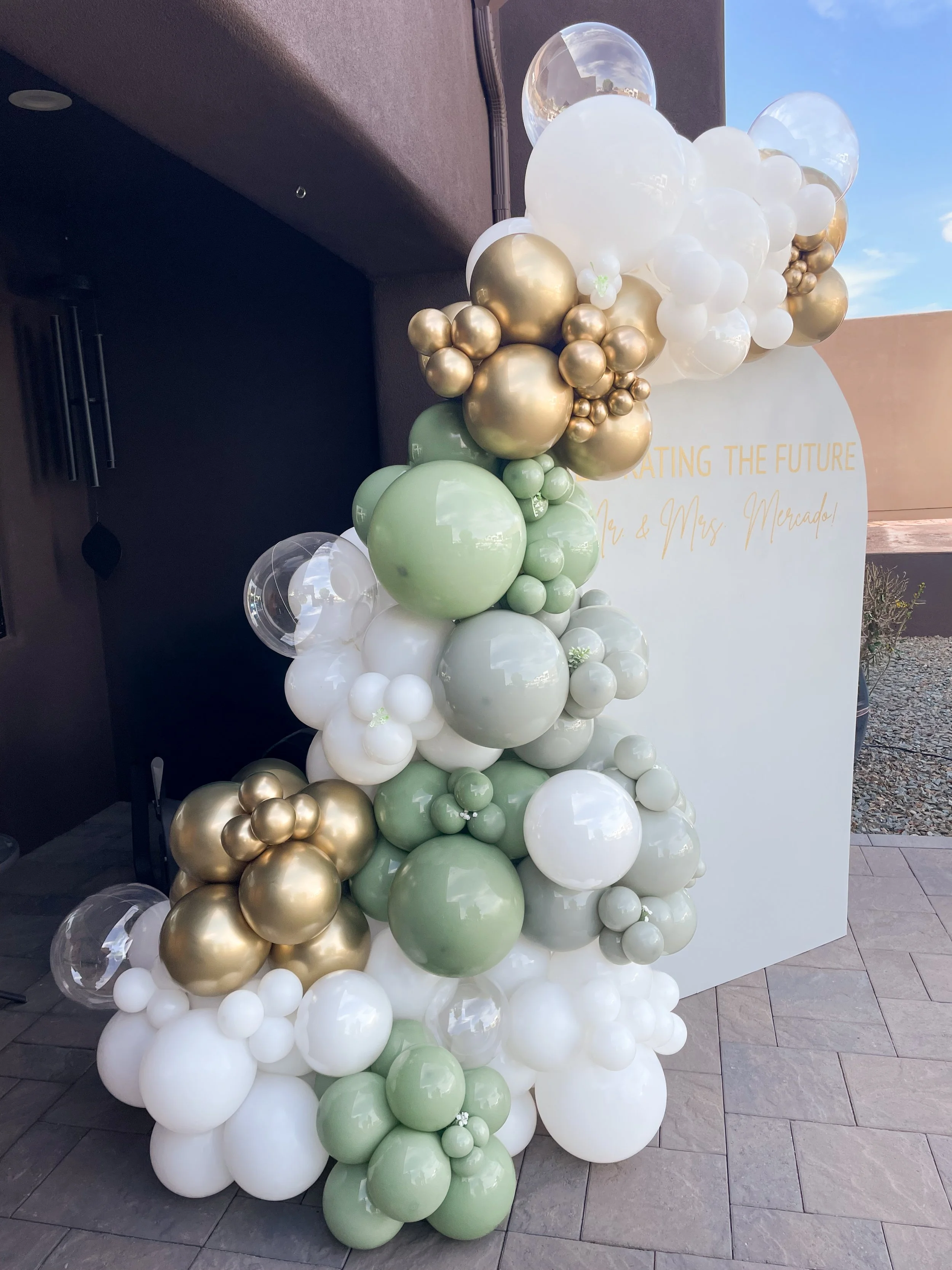 A custom backdrop with organic balloon garland in white, gold, and sage green balloons with gold writing. Engagement Party Decor