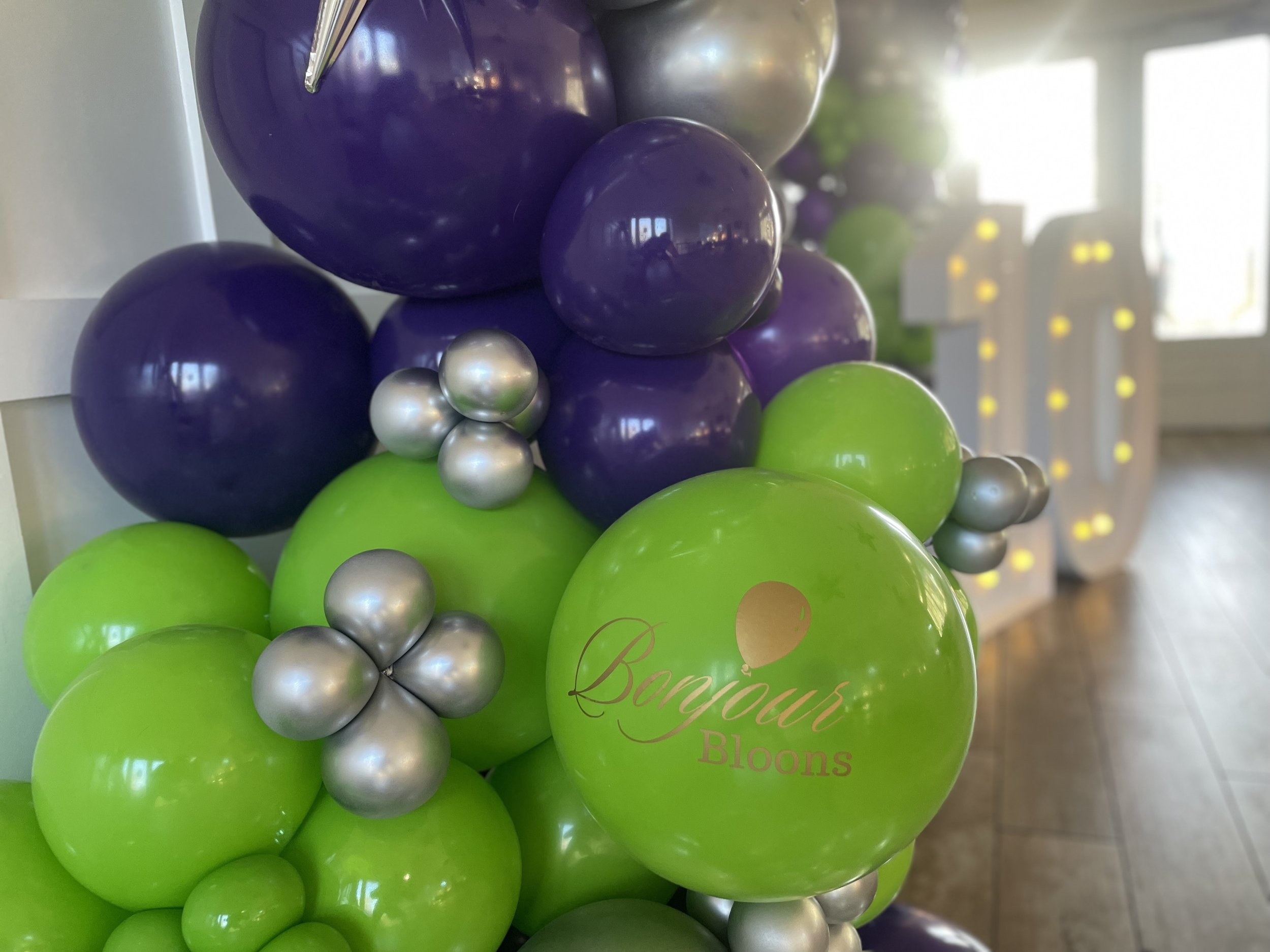 A cluster of balloons in purple, silver, and bright green colors, with the green balloon having the words 'Bonjour Bloons' written on it.