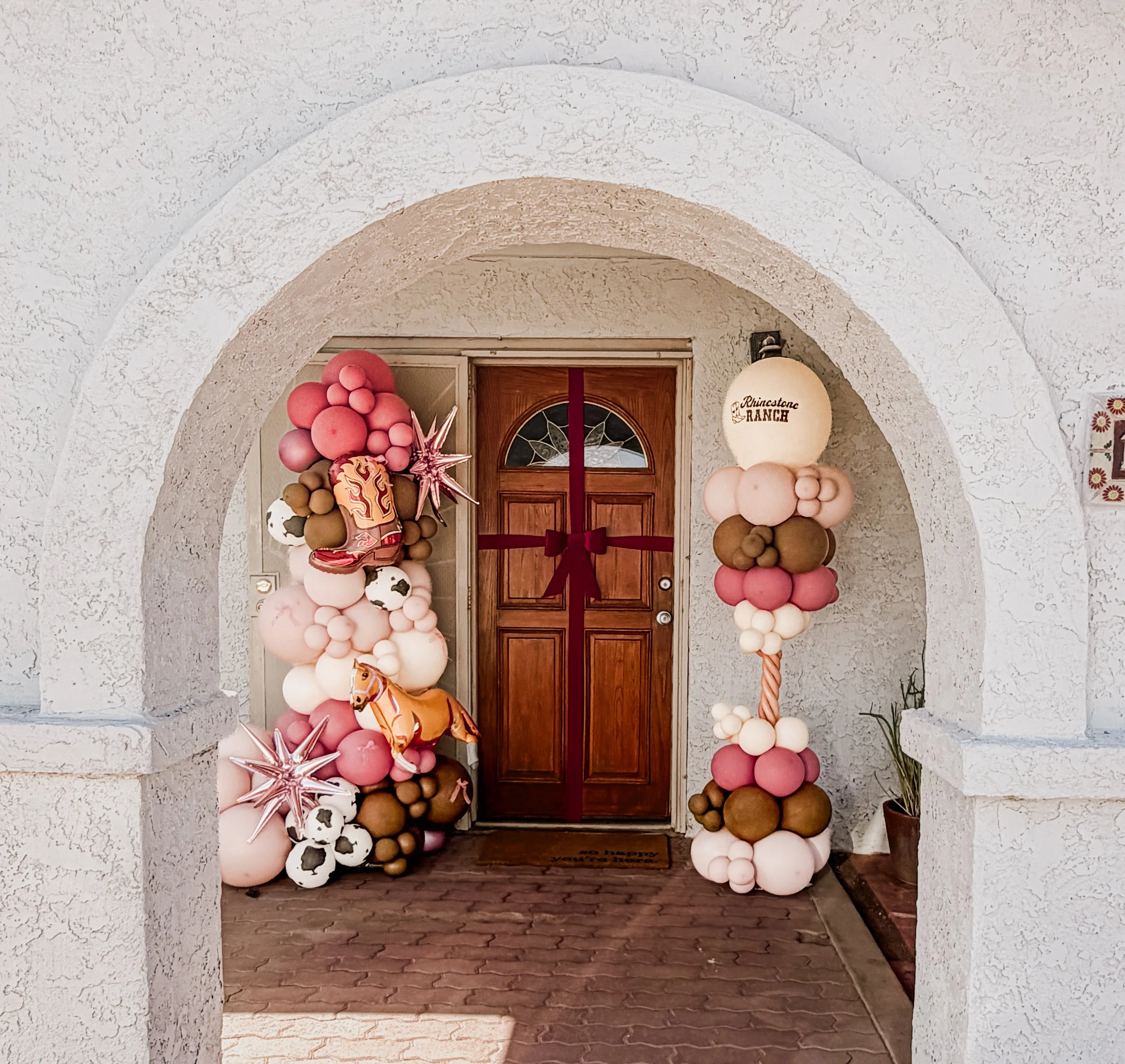 Decorative freestanding cowgirl themed balloon garland in pink, white, and brown colors, including star-shaped balloons, framing a wooden door.