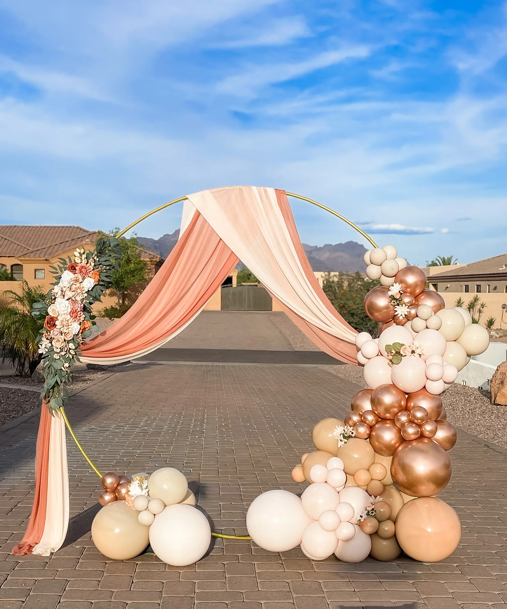 A circle hoop  white, rose gold, and peach balloons arranged in a cascading design. The backdrop includes a peach and pink fabric drape, flowers, and greenery, set outdoors with a mountain range, houses, and a blue sky with clouds. Vow Renewal Backdr