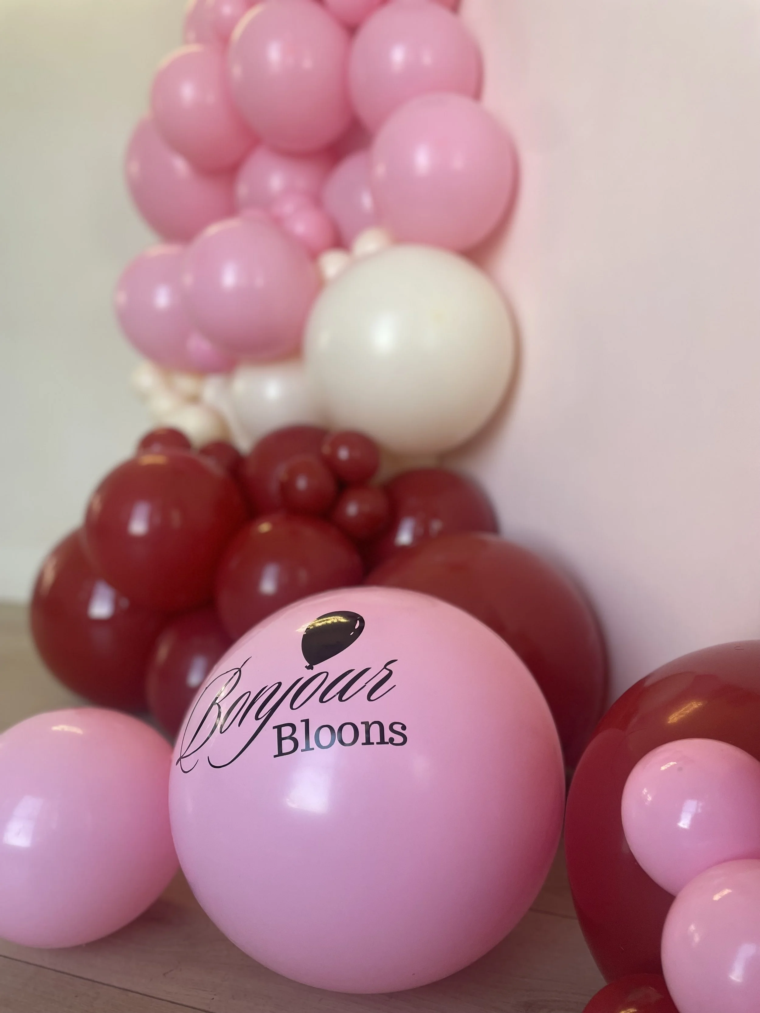 Pink and red balloons arranged in a cluster with a pink balloon featuring black cursive text saying 'Bonjour Bloons'