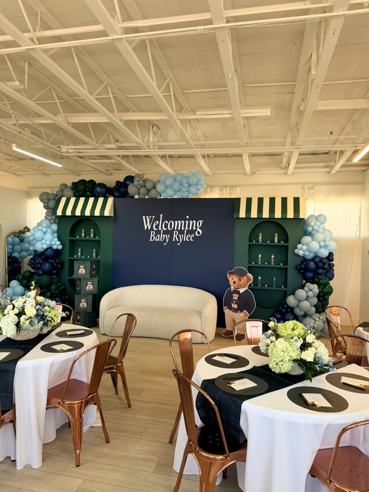 Baby shower celebration setup with a decorated backdrop saying "Welcoming Baby Rylee" featuring balloons in shades of blue, white, and green. There are round tables with white tablecloths, black accents, floral centerpieces, golden chairs, and place 