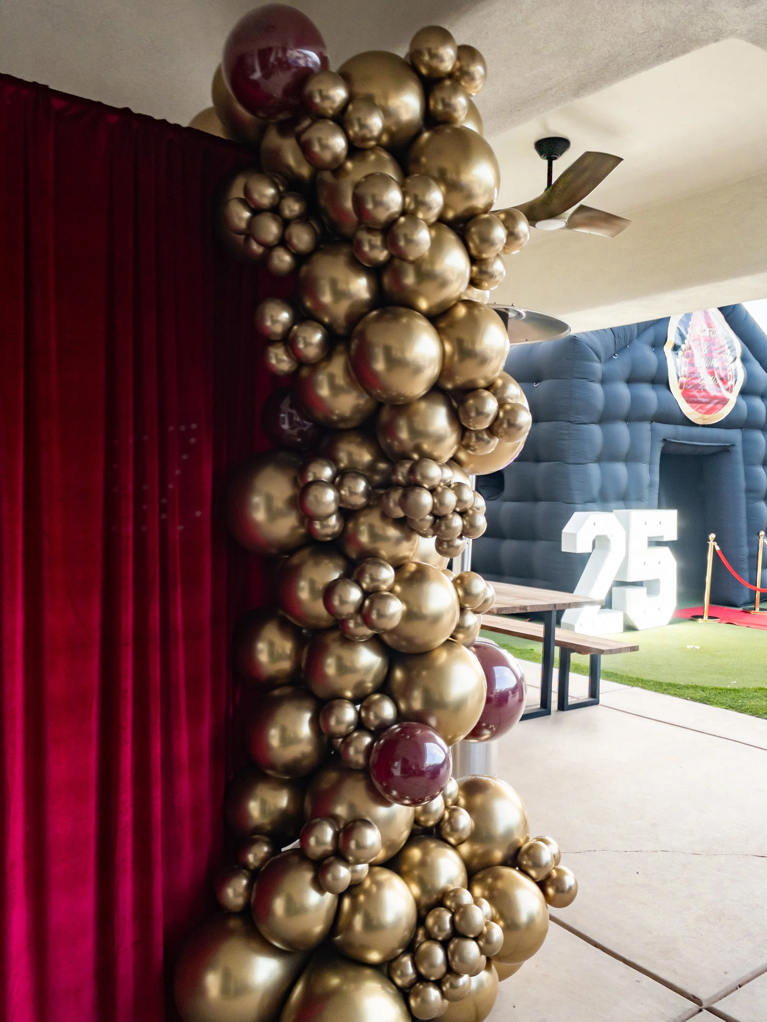 gold and maroon organic balloons next to red velvet curtain, part of a 25th celebration with large white '25' numbers and red carpet, inside a venue.