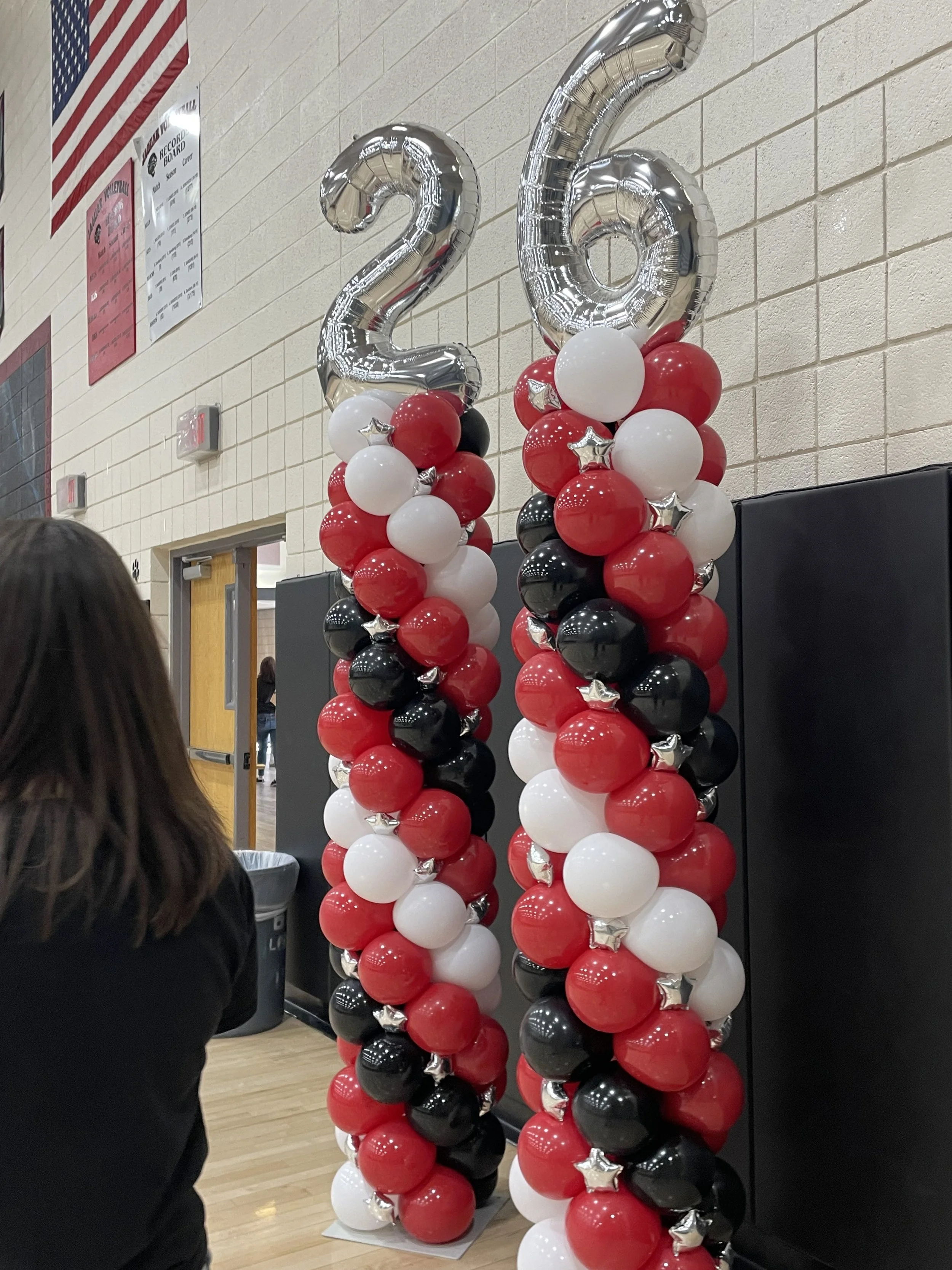 Two balloon columns decorated with red, black, and white balloons topped with large silver number balloons '2' and '6', standing on a gymnasium floor.