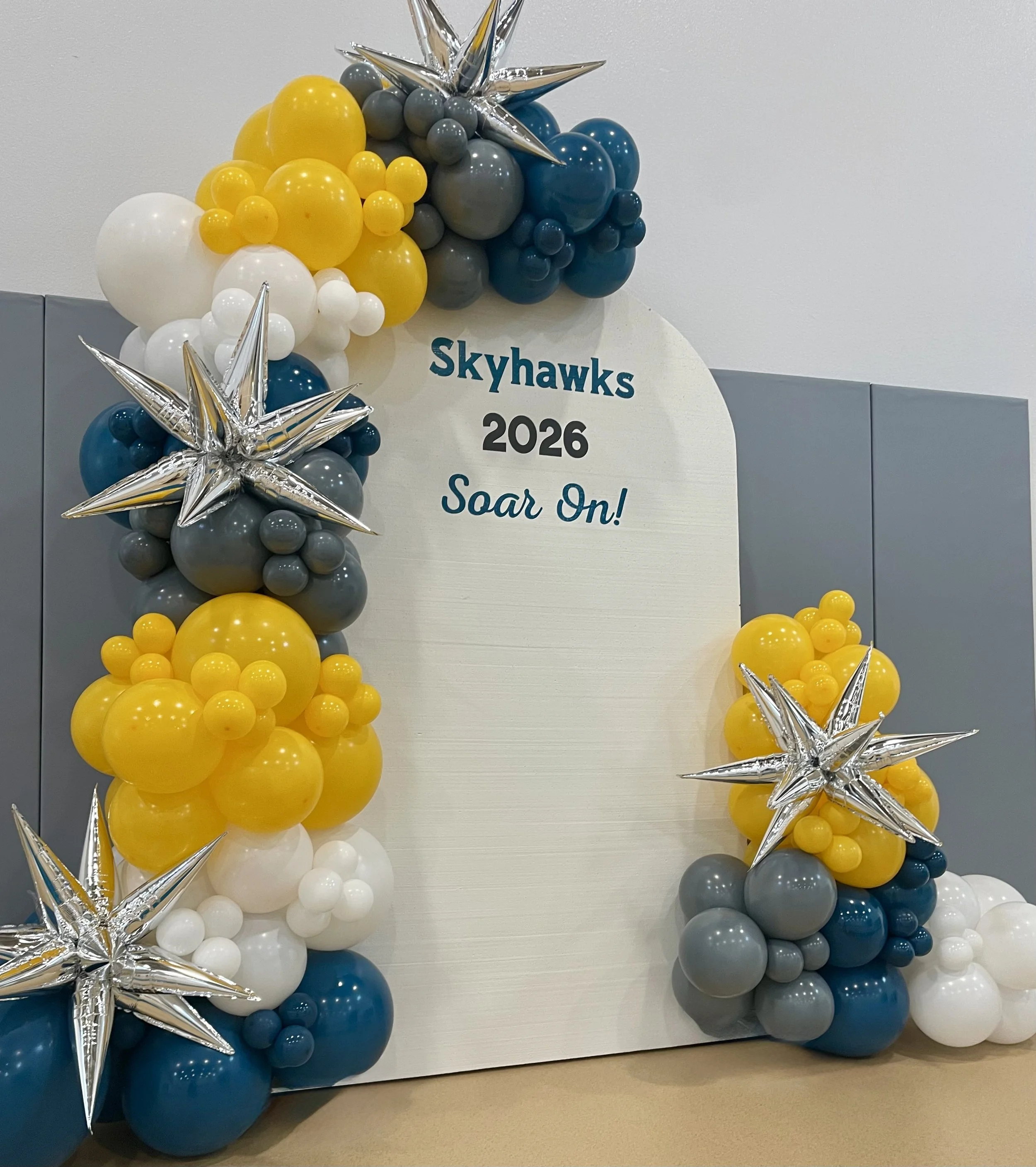 Balloon arrangement with yellow, white, gray, and blue balloons forming a decorative display for Skyhawks 2026 event, accented with silver star-shaped balloons.