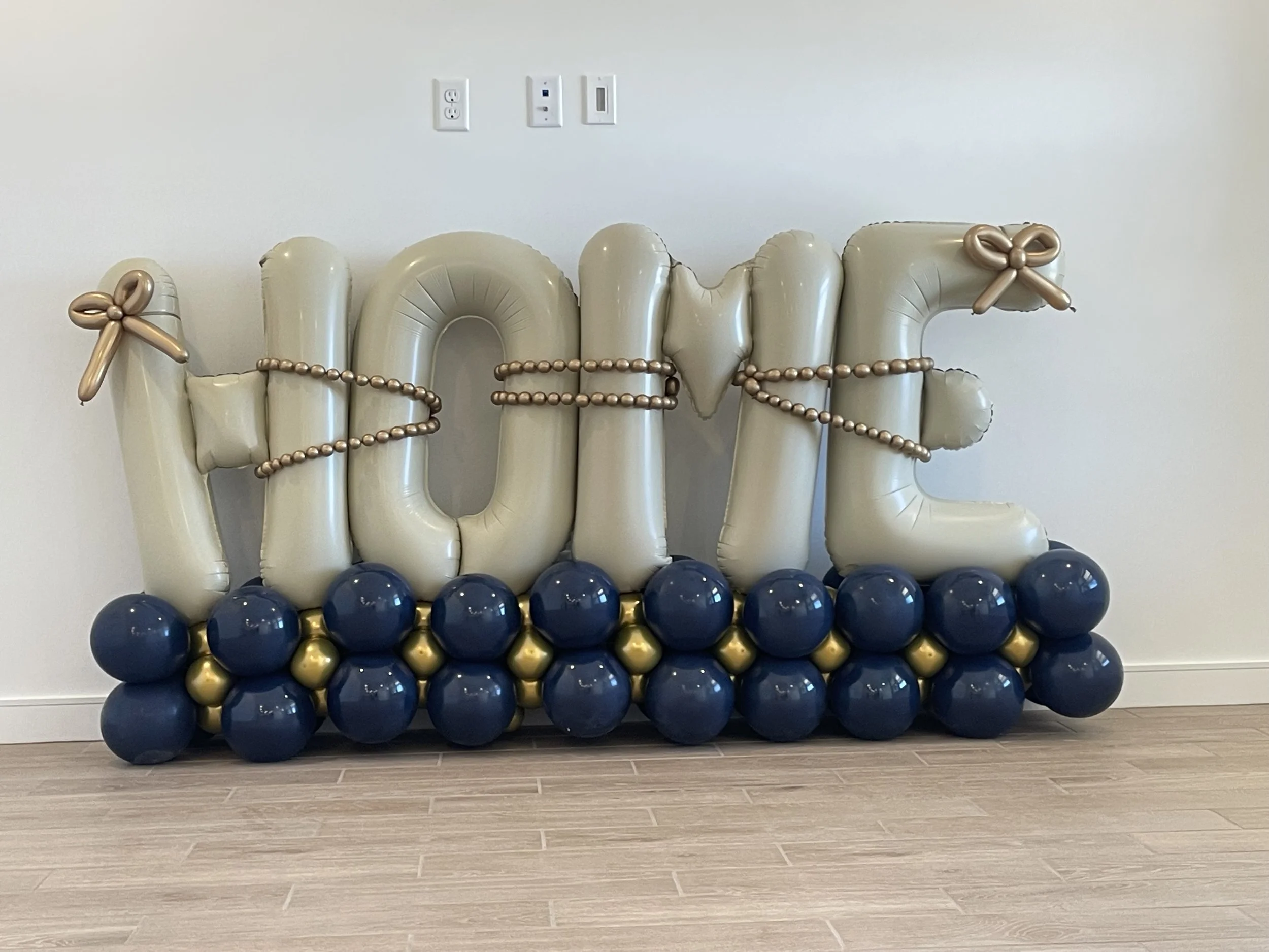Balloon decorations spelling out 'home' with gold and navy blue balloons at the bottom, gold balloon bows on the letters, and a string of gold beads wrapping around the balloons. Housewarming - Balloon Stack
