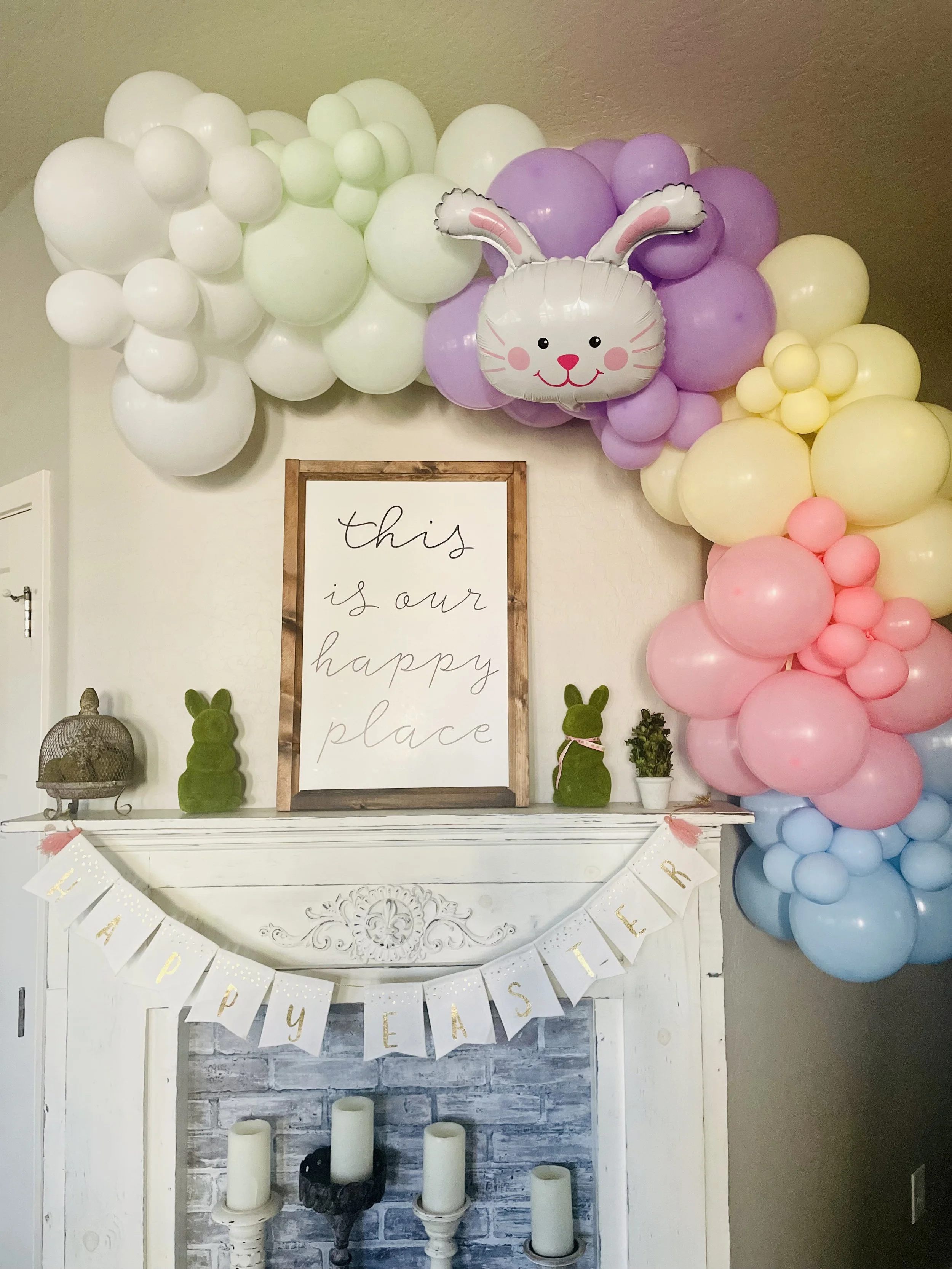 Decorative display with pastel-colored balloons arranged in clusters, including white, purple, pink, yellow, and blue, along with bunny-shaped foil balloons. A sign on the fireplace mantel reads 'this is our happy place'. A banner hanging beneath spe