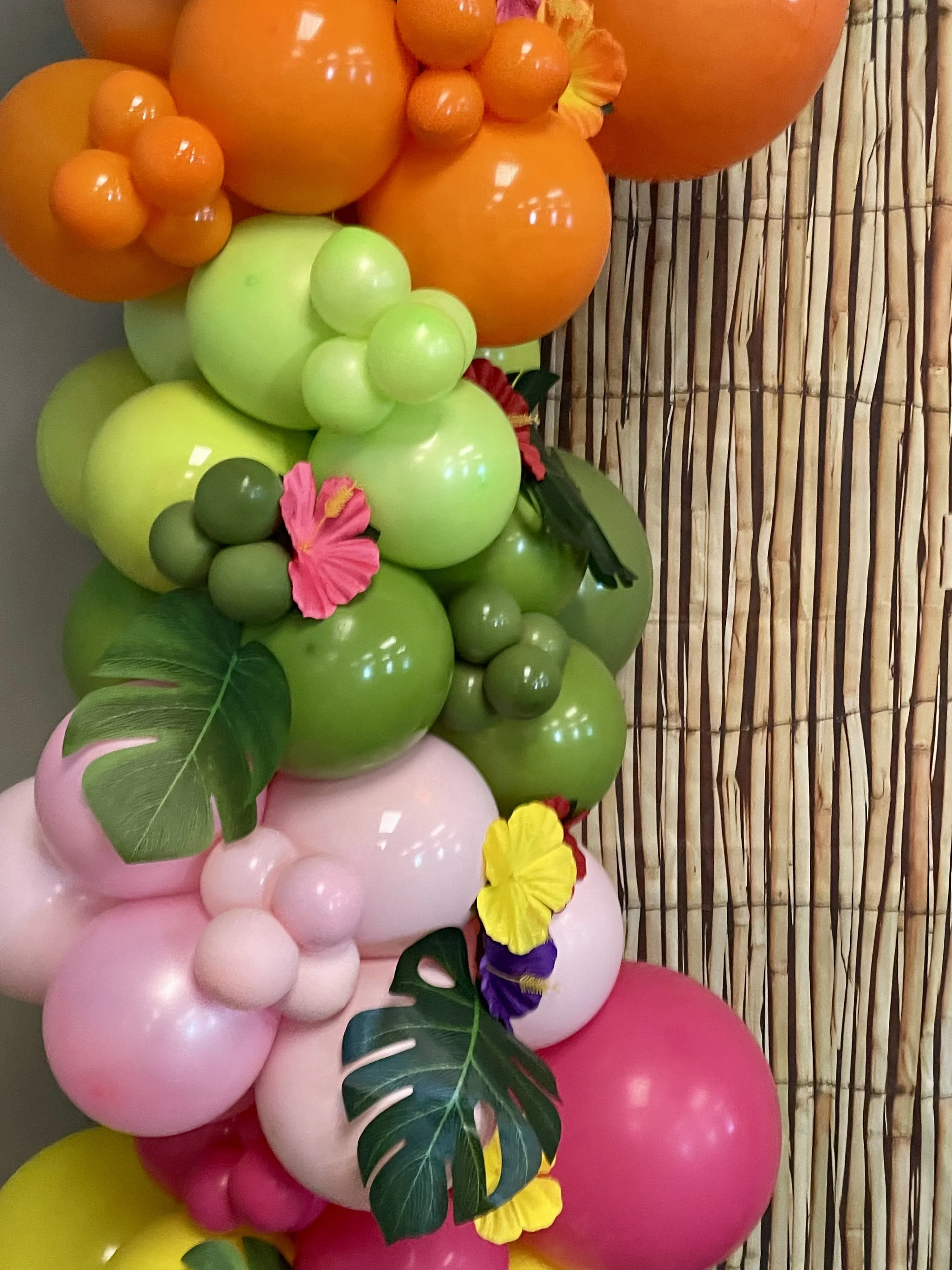 Colorful balloon display with pink, green, yellow, and orange balloons, decorated with tropical leaves and flowers, with a bamboo background.