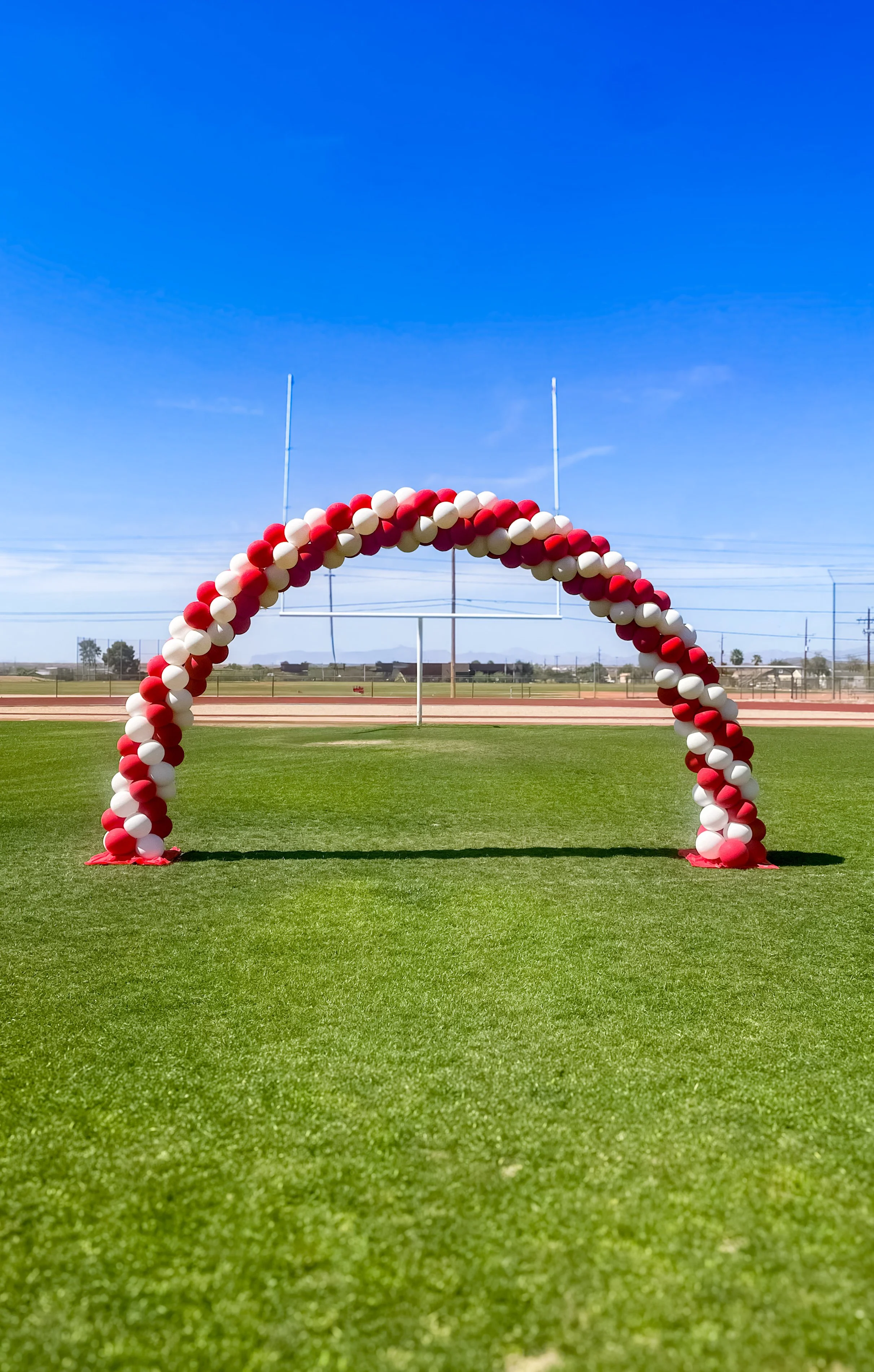 A celebration arch made of red and white balloons on a sports field with goalposts, under a clear blue sky.