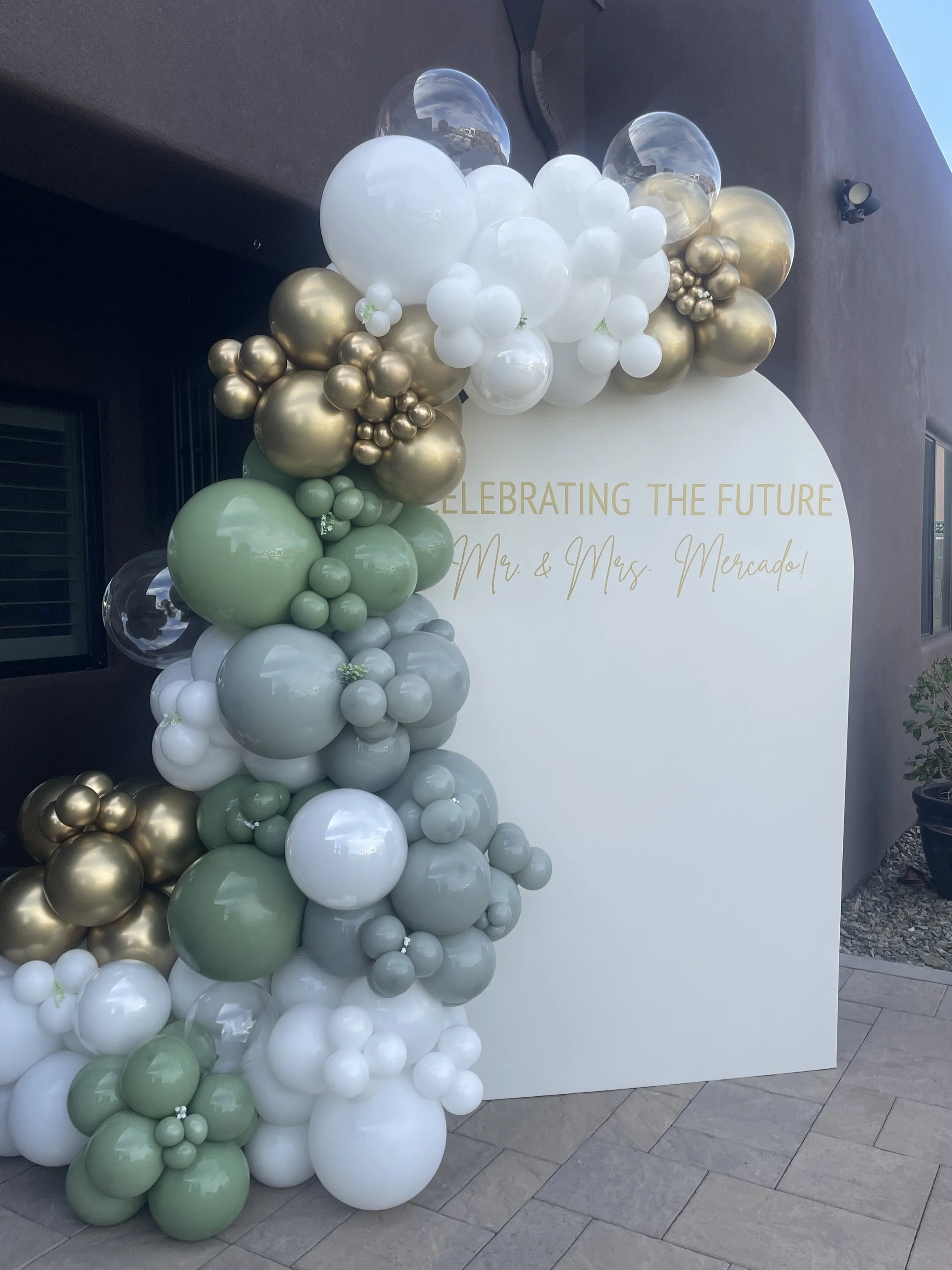 Balloon arch with white, gold, and light green balloons, and a white backdrop with gold text that reads 'Celebrating the Future Mr. & Mrs. Mercado'.